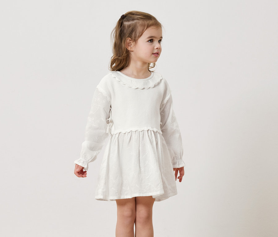 Mädchen mit blonden Haaren trägt ein weißes, langärmeliges Kleid vor einem neutralen Hintergrund.