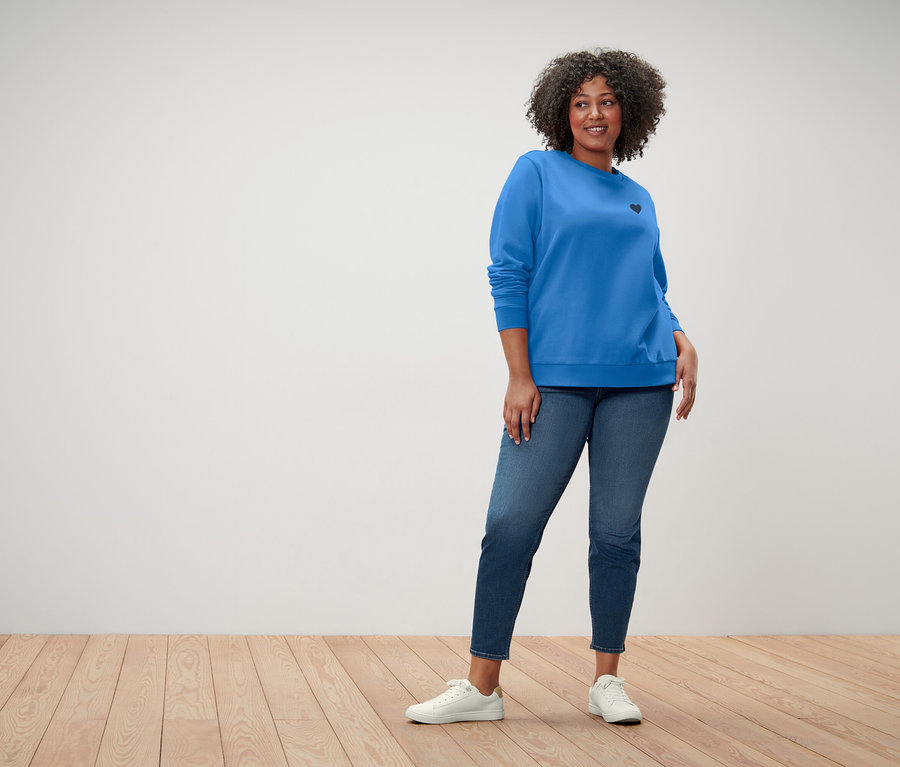 Frau mit lockigem Haar trägt einen blauen Pullover mit einem kleinen schwarzen Herz, blaue Jeans und weiße Turnschuhe.