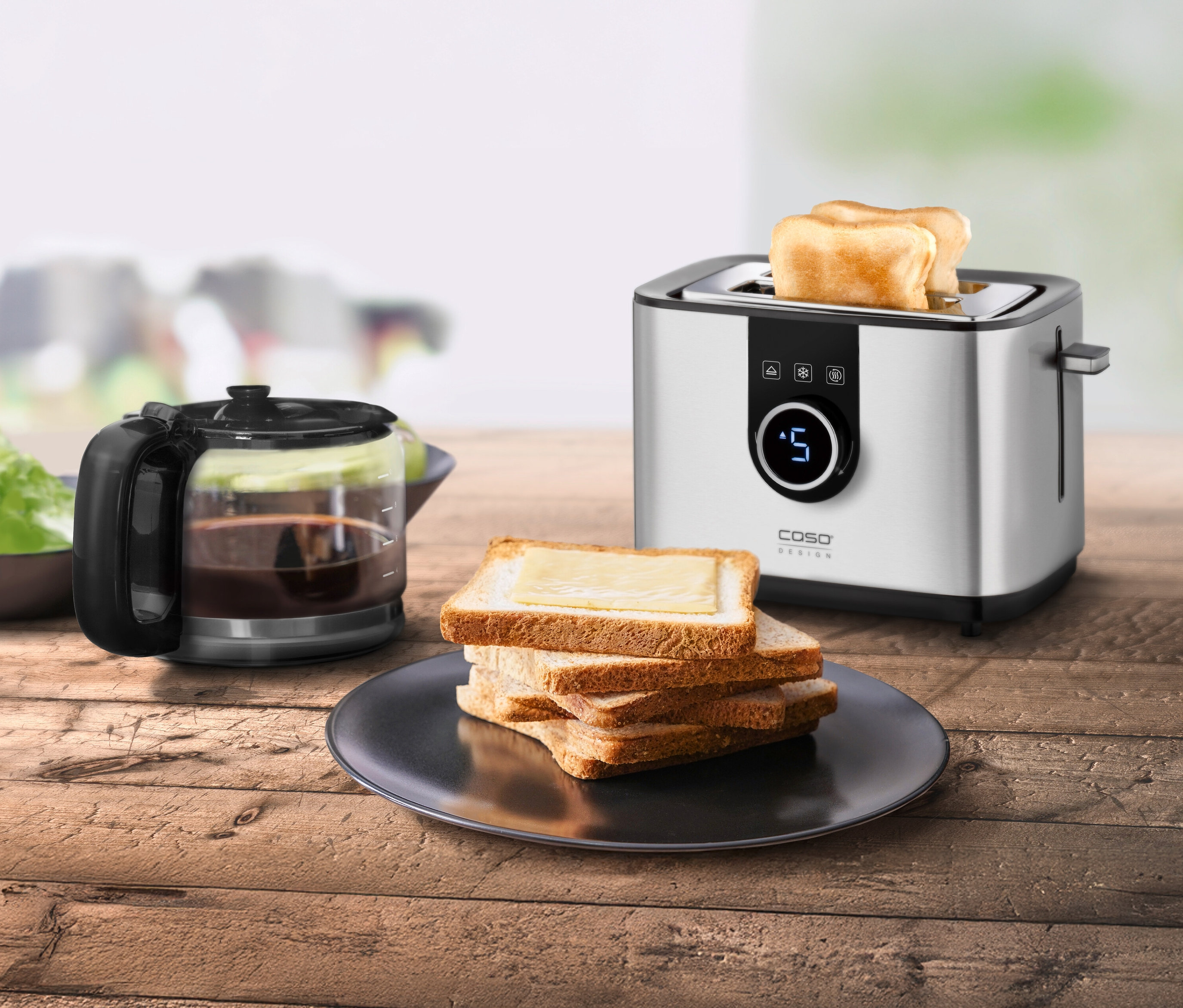 Ein silberner Toaster Selection T2 steht neben einem Stapel Toastbrot und einer Kaffeekanne auf einem Holztisch.
