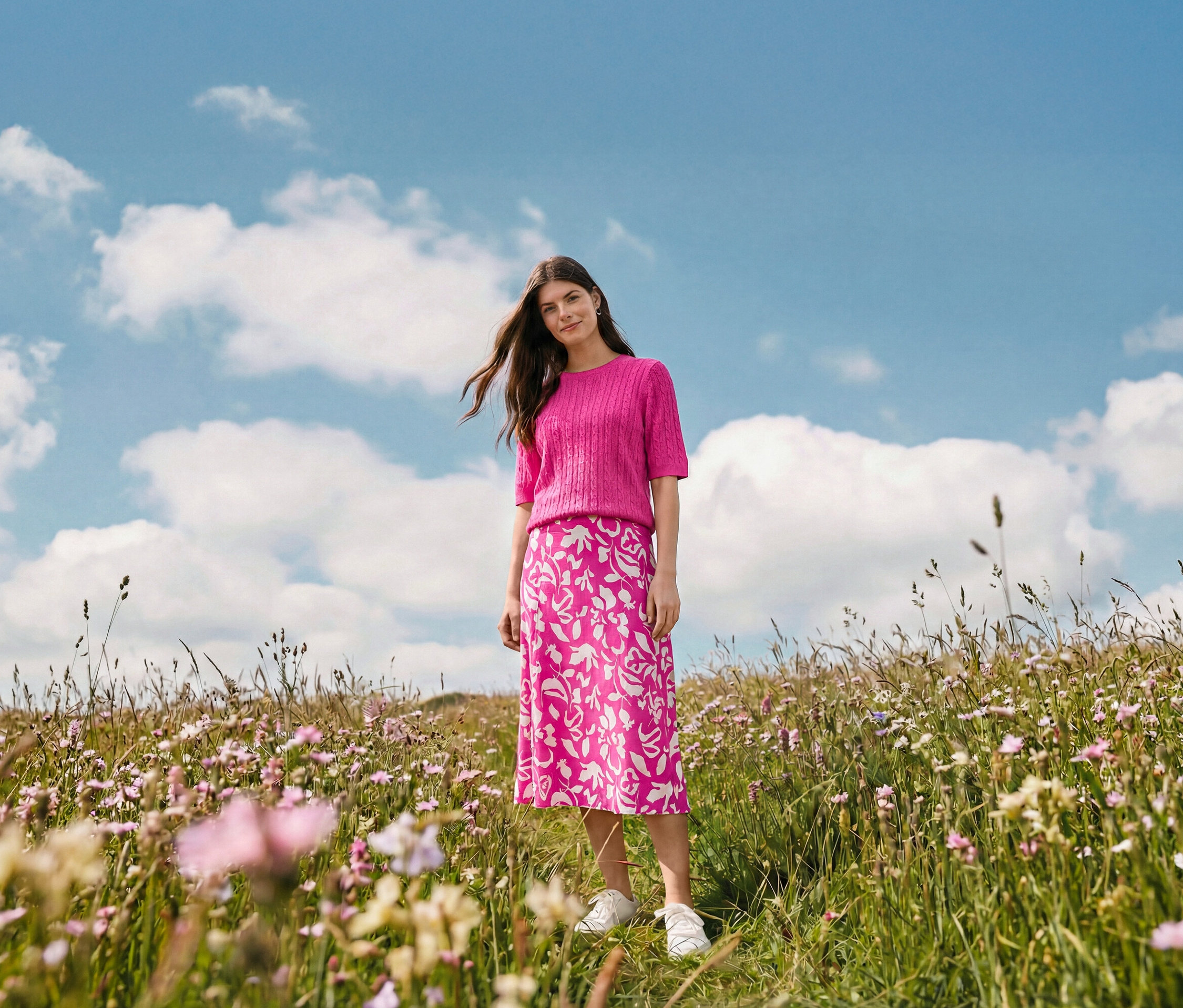 Eine Frau steht auf einer Wiese und trägt einen bedruckten Midi-Rock, Creolen Wave und Leder-Sneaker.