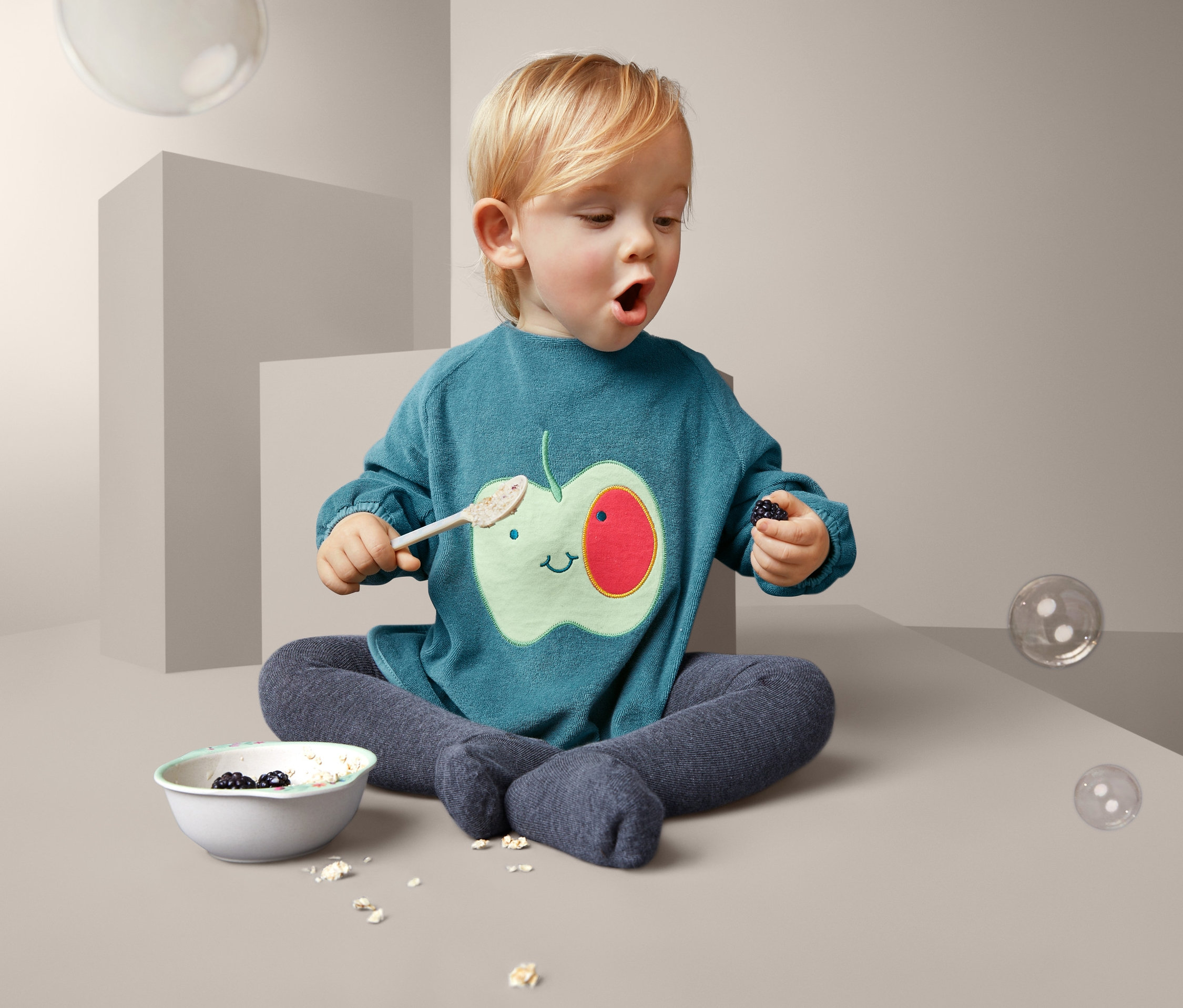 Kleinkind sitzt mit Schüssel und Löffel und trägt ein Lätzchen mit Apfel.