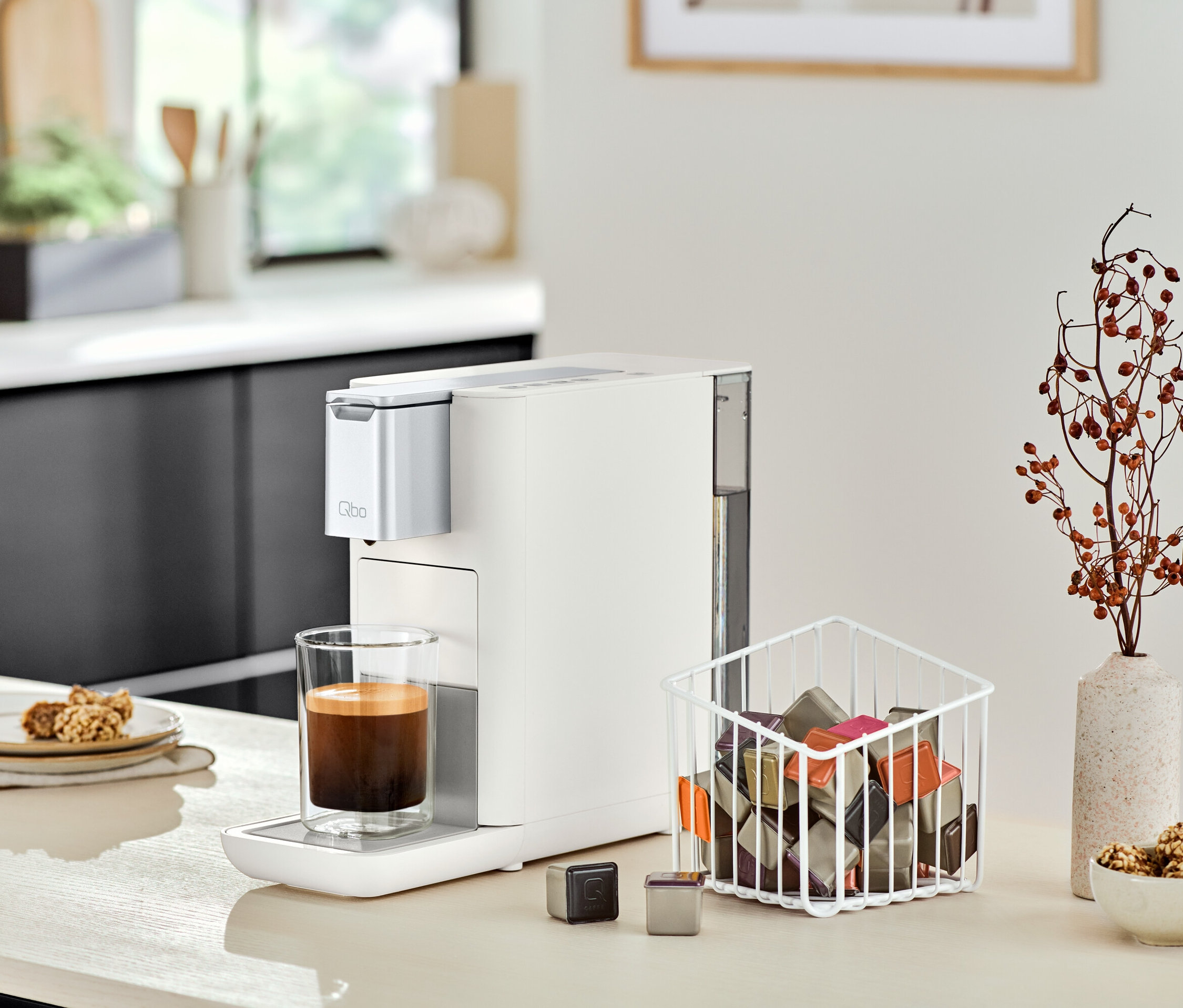 Eine Qbo ESSENTIAL Bright Sand Kaffeemaschine steht neben einem Glas Kaffee auf einer Küchentheke.