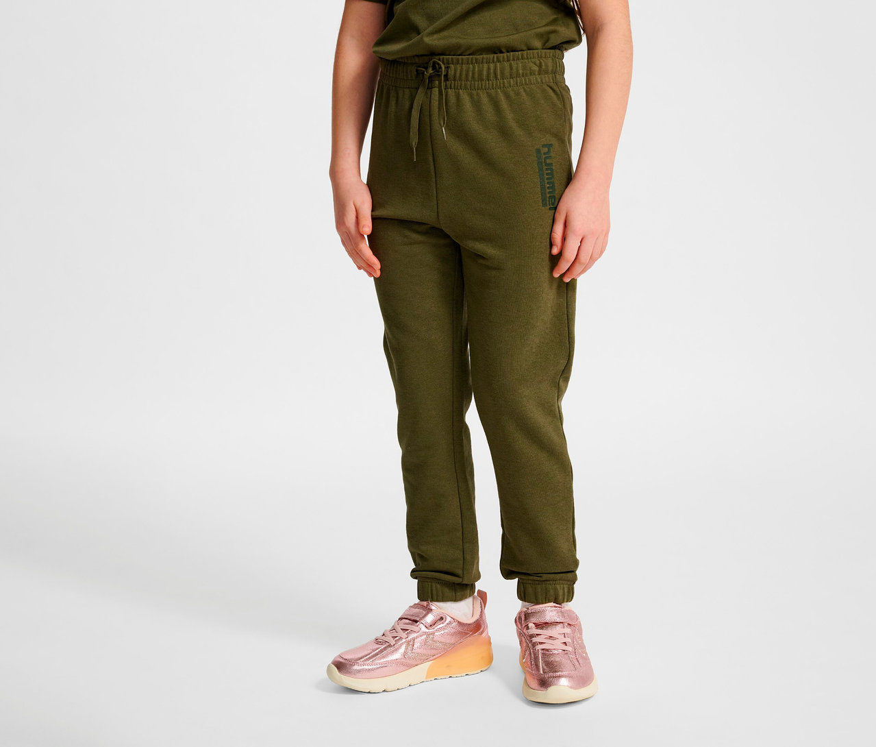 Eine Person steht und trägt eine grüne HUMMEL HMLJR Base Regular Pants und rosa Turnschuhe.
