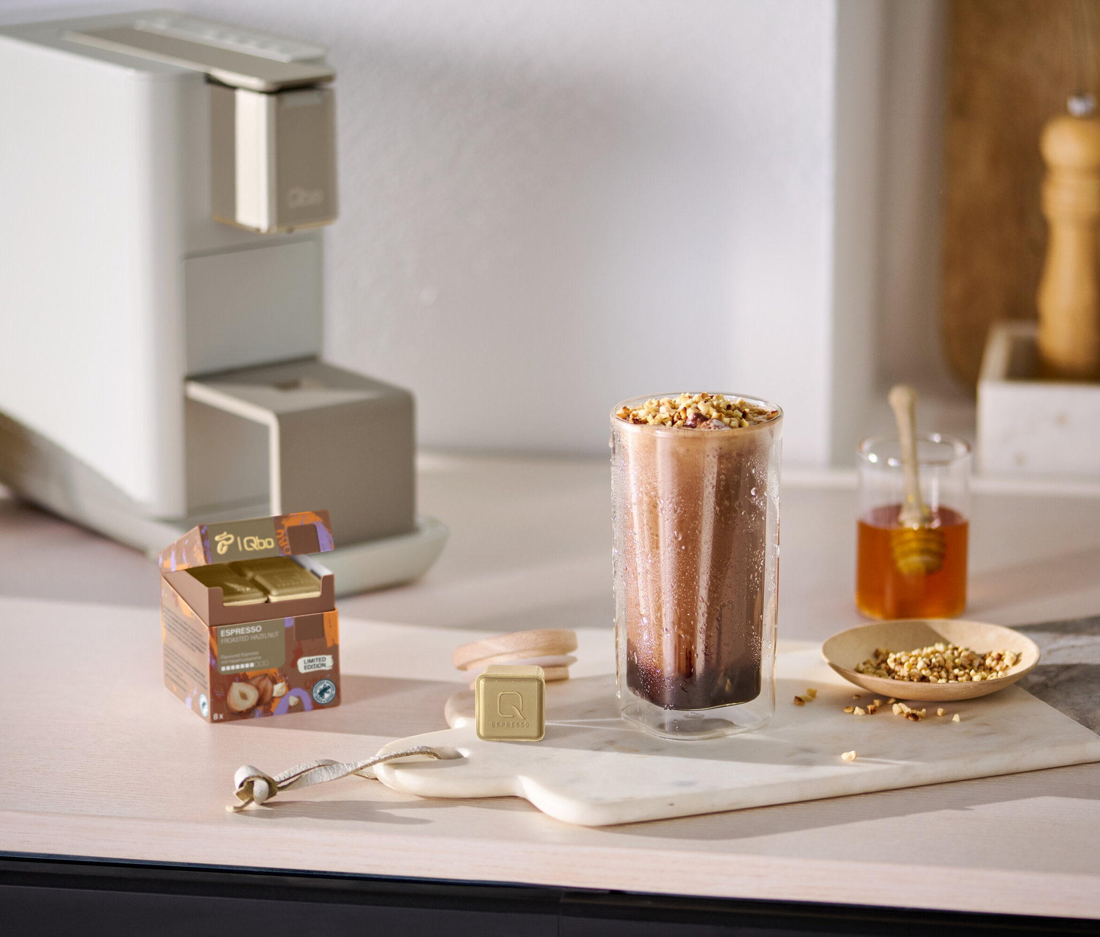 Auf einem Tisch stehen eine Qbo ICONIC Dark Chrome Kaffeemaschine, ein Glas mit einem Getränk, Honig und Nüsse.