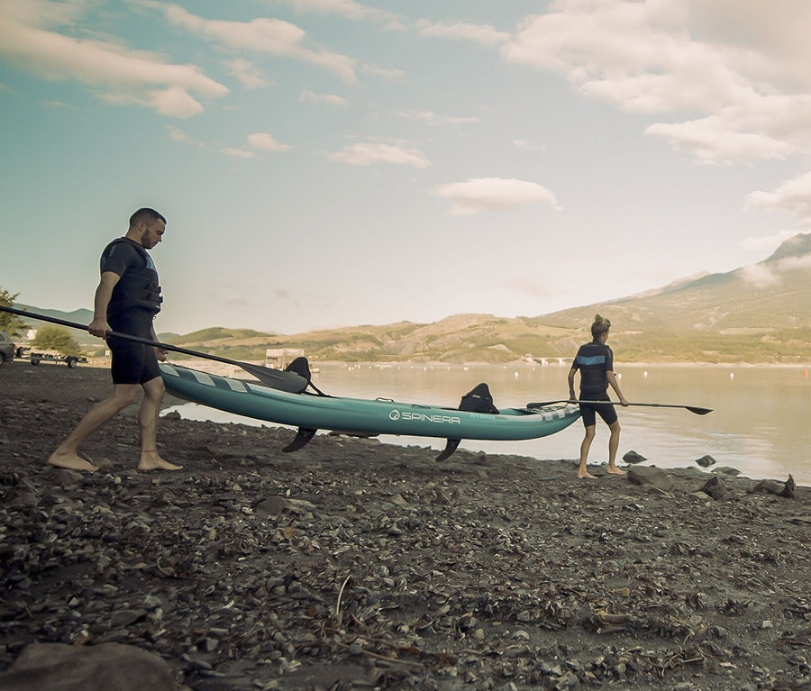 Zwei Personen tragen ein Spinera Hybris 410 2-Personen-Kajak am Ufer eines Sees.