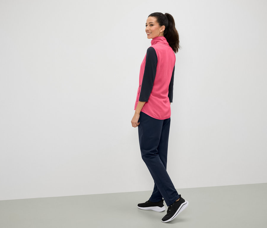 Eine Frau lächelt über die Schulter, sie trägt eine pinke Weste, marineblaue Hosen und schwarze Turnschuhe.