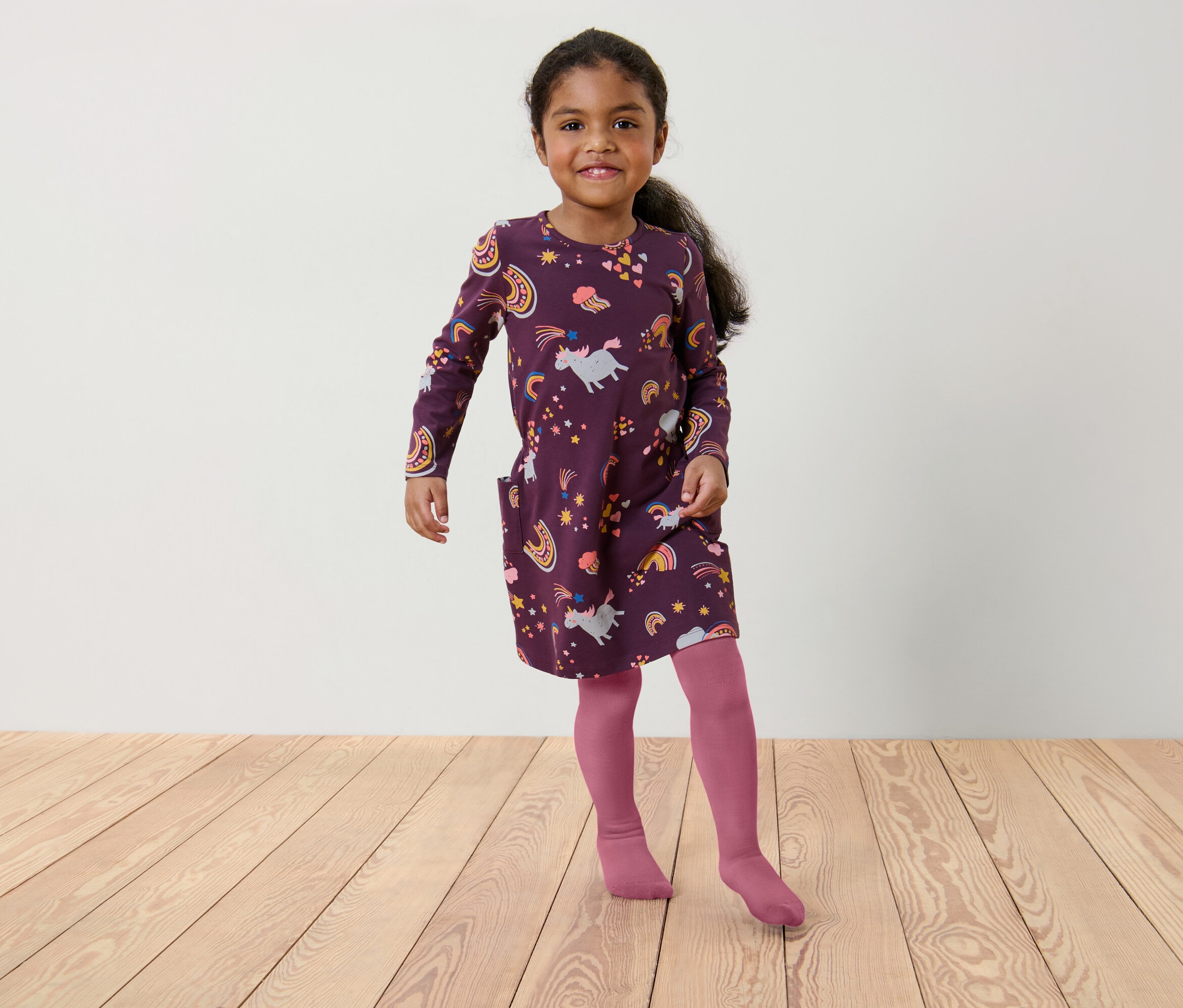 Mädchen in einem burgunderroten Kleid mit Einhorn- und Regenbogenmuster und rosa Strumpfhosen steht auf einem Holzboden.