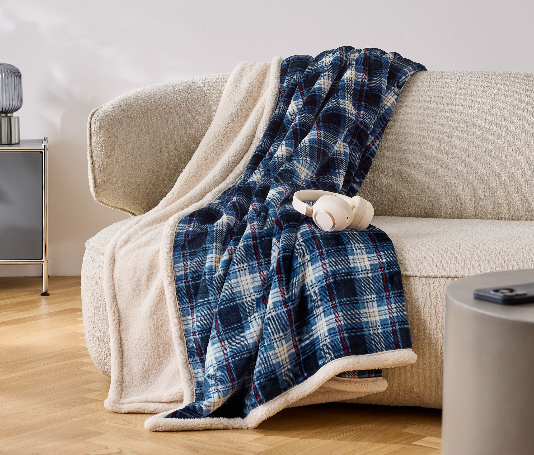 Auf einem beigen Sofa liegt eine blau-weiss karierte Decke mit weissem Rand. Auf der Decke liegen weisse Kopfhörer.