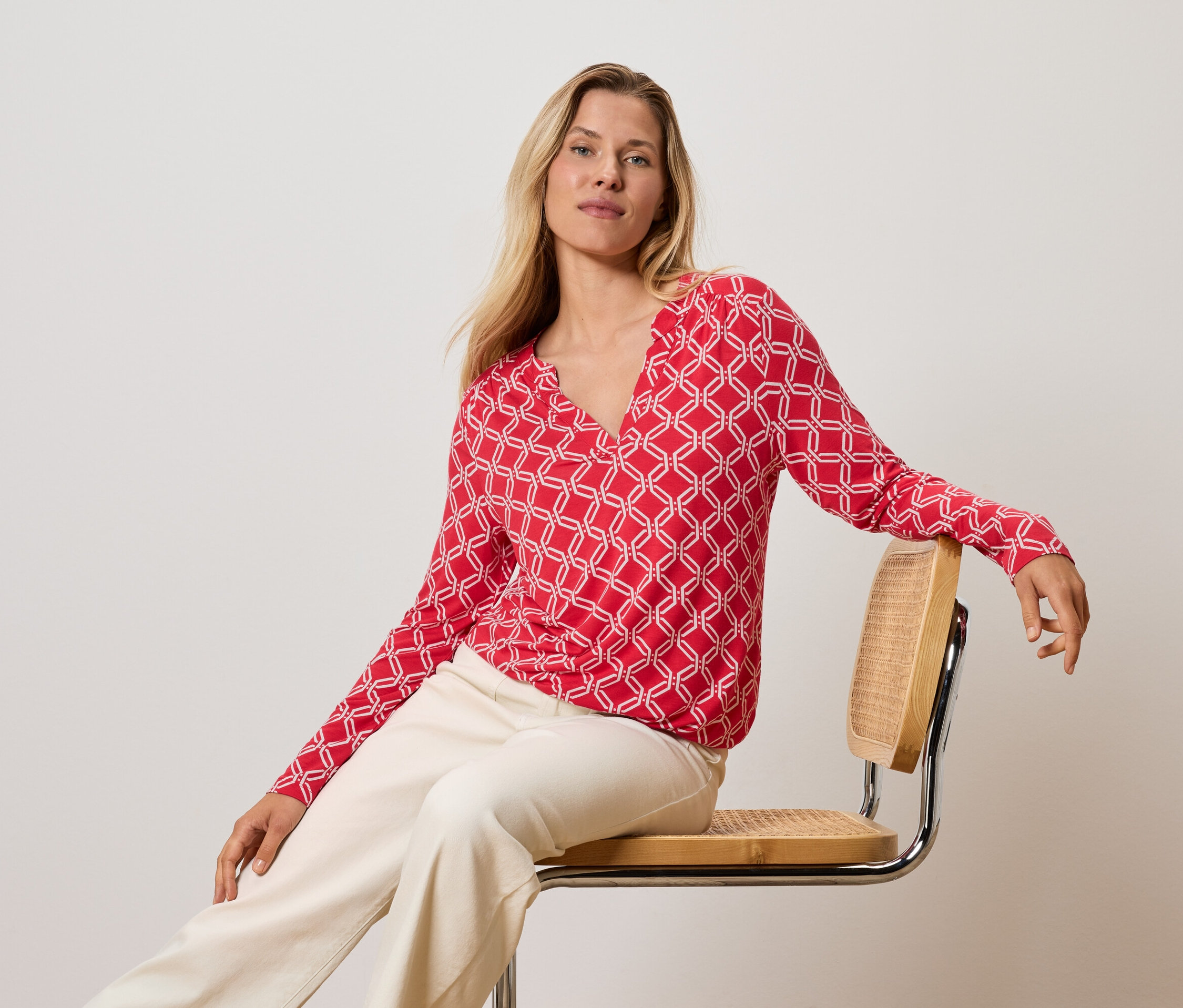 Blonde Frau sitzt auf einem Stuhl, trägt ein rot-weißes Oberteil und cremefarbene Hosen.