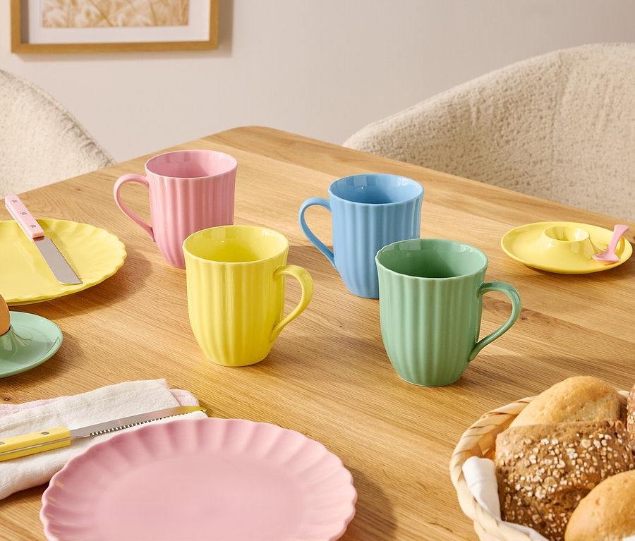 Auf einem Holztisch stehen Teller und Tassen in Pastellfarben sowie ein Brotkorb.