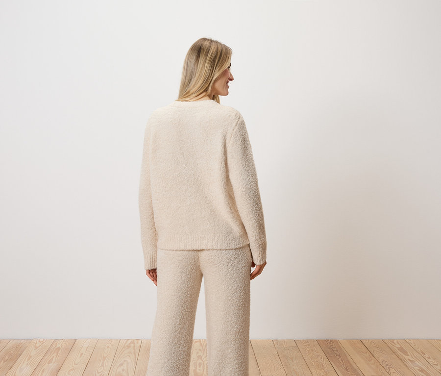 Eine Frau mit blonden Haaren steht mit dem Rücken zur Kamera und trägt einen cremefarbenen Pullover und Hose.