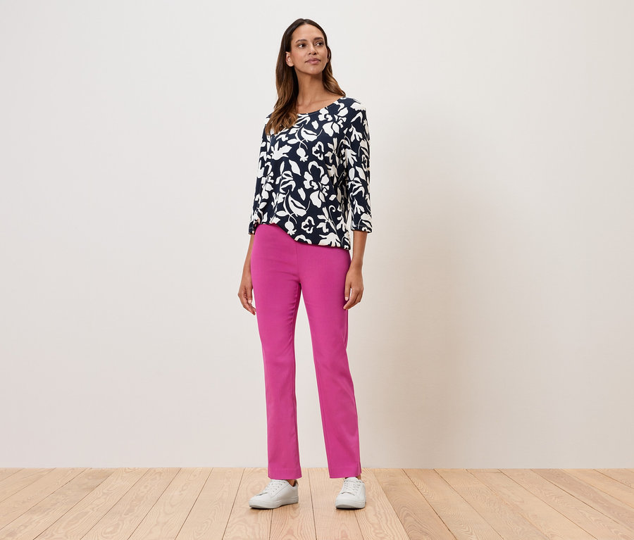 Eine Frau steht und trägt ein geblümtes Oberteil mit Dreiviertelärmeln, eine pinke Hose und weiße Turnschuhe.