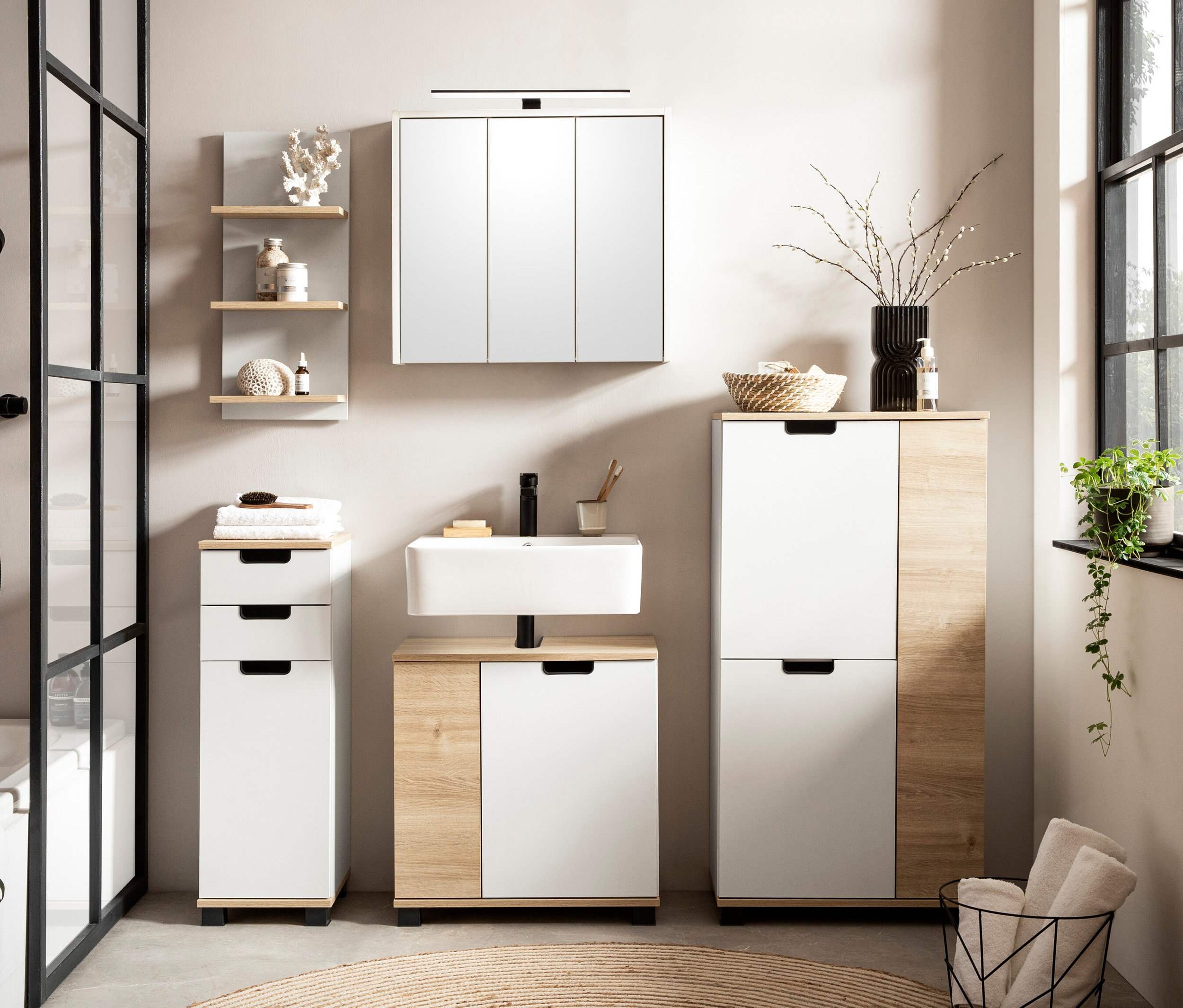 Badezimmer-Interieur mit Waschbecken, Spiegelschrank, Waschbeckenunterschrank »Emmik« in Weiß/Weißeiche, Highboard »Emmik« in Weiß/Weißeiche und Regal »Emmik« in Weiß/Weißeiche.