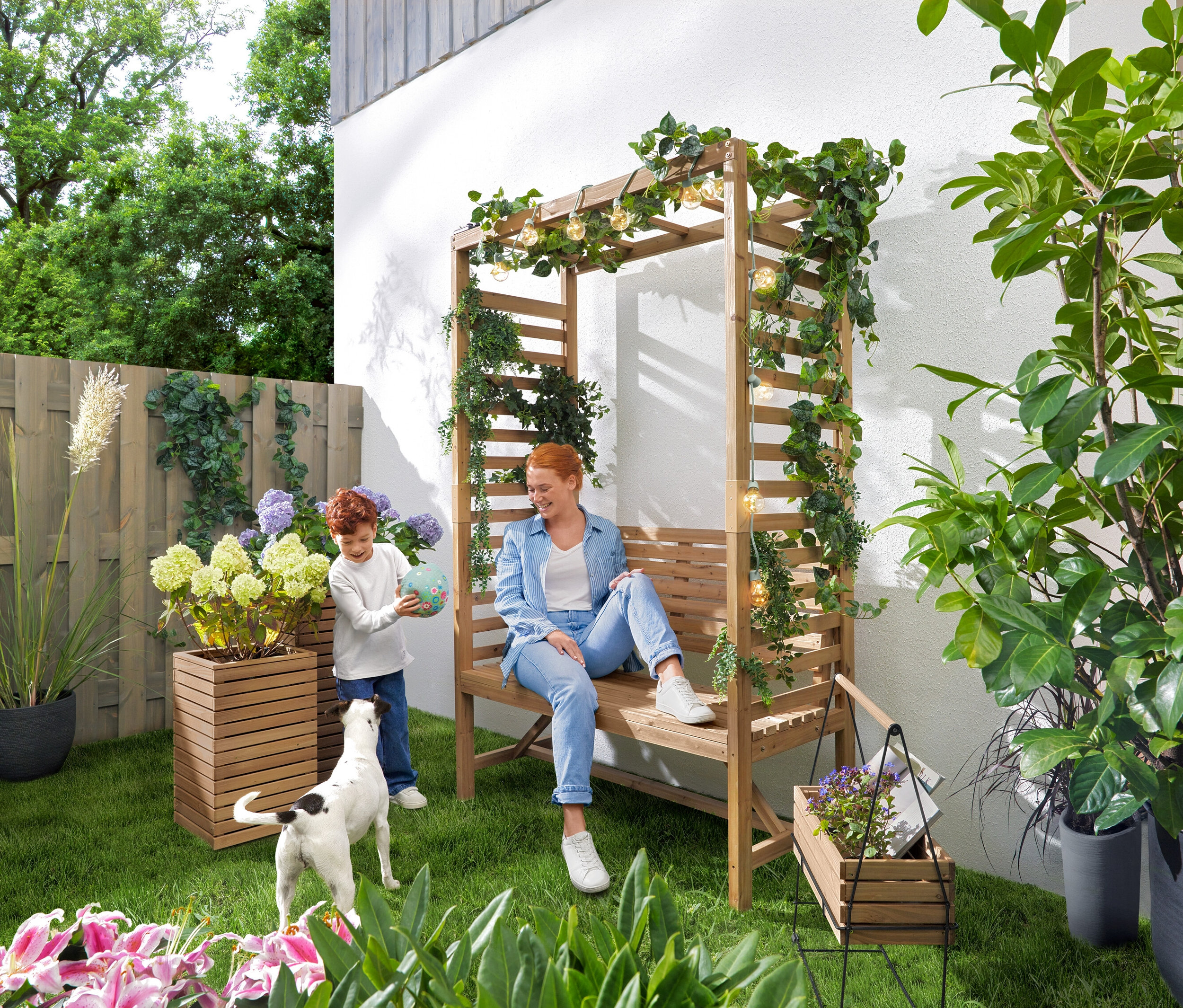 Eine Frau sitzt auf einer Gartenbank mit Rosenbogen, während ein Kind mit einem Ball spielt. Eine Solar-Lichterkette »Glühbirnen« hängt über der Bank. Im Garten stehen 2 Pflanzsäulen »Tinus« und ein Kräuterkasten mit Tragegestell »Elin«.