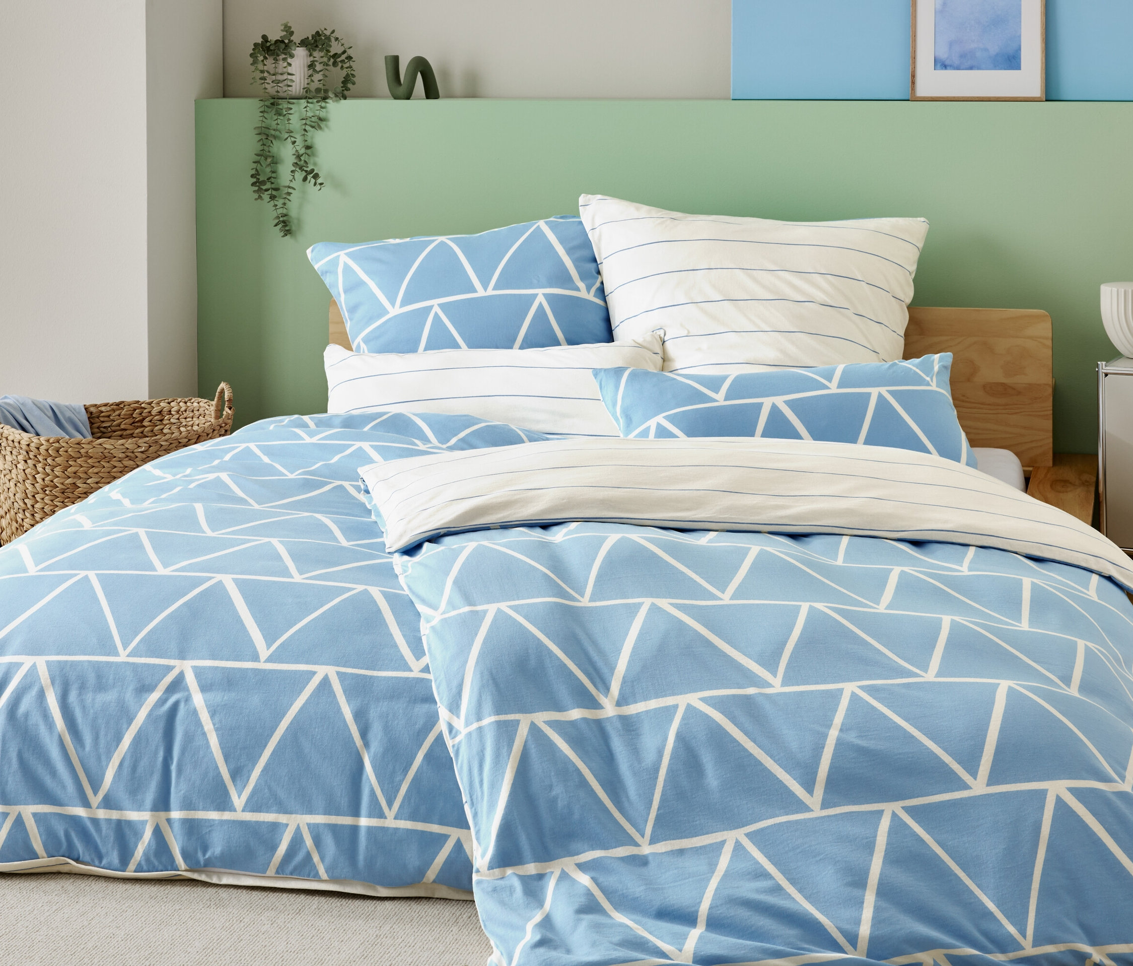 Bett mit hellblauem Bezug mit weißen Dreiecken und gestreiften Kissenbezügen, neben einem Korbstuhl und einem Nachttisch mit Lampe.