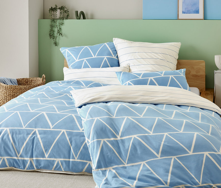 Bett mit hellblauem Bezug mit weißen Dreiecken und gestreiften Kissenbezügen, neben einem Korbstuhl und einem Nachttisch mit Lampe.