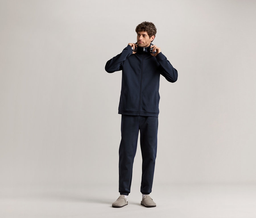 Mann steht und hält Kopfhörer in der Hand, trägt einen dunkelblauen Hausanzug.