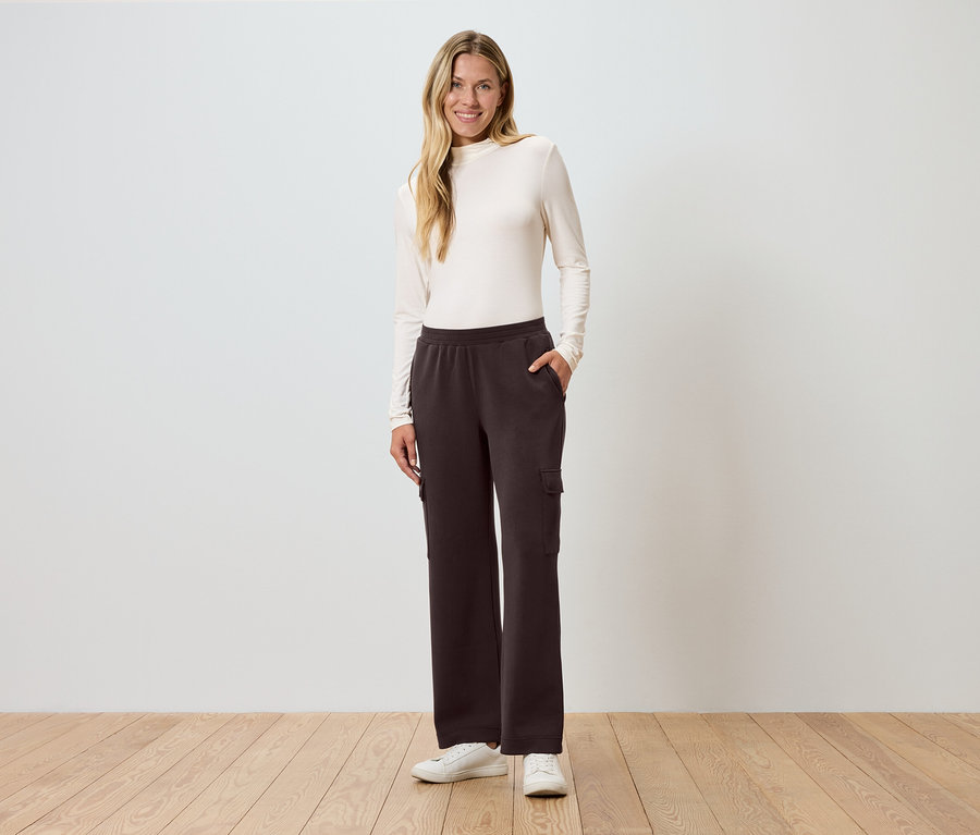 Frau steht mit Hand in Tasche vor einer Wand und trägt braune Cargo-Hose.