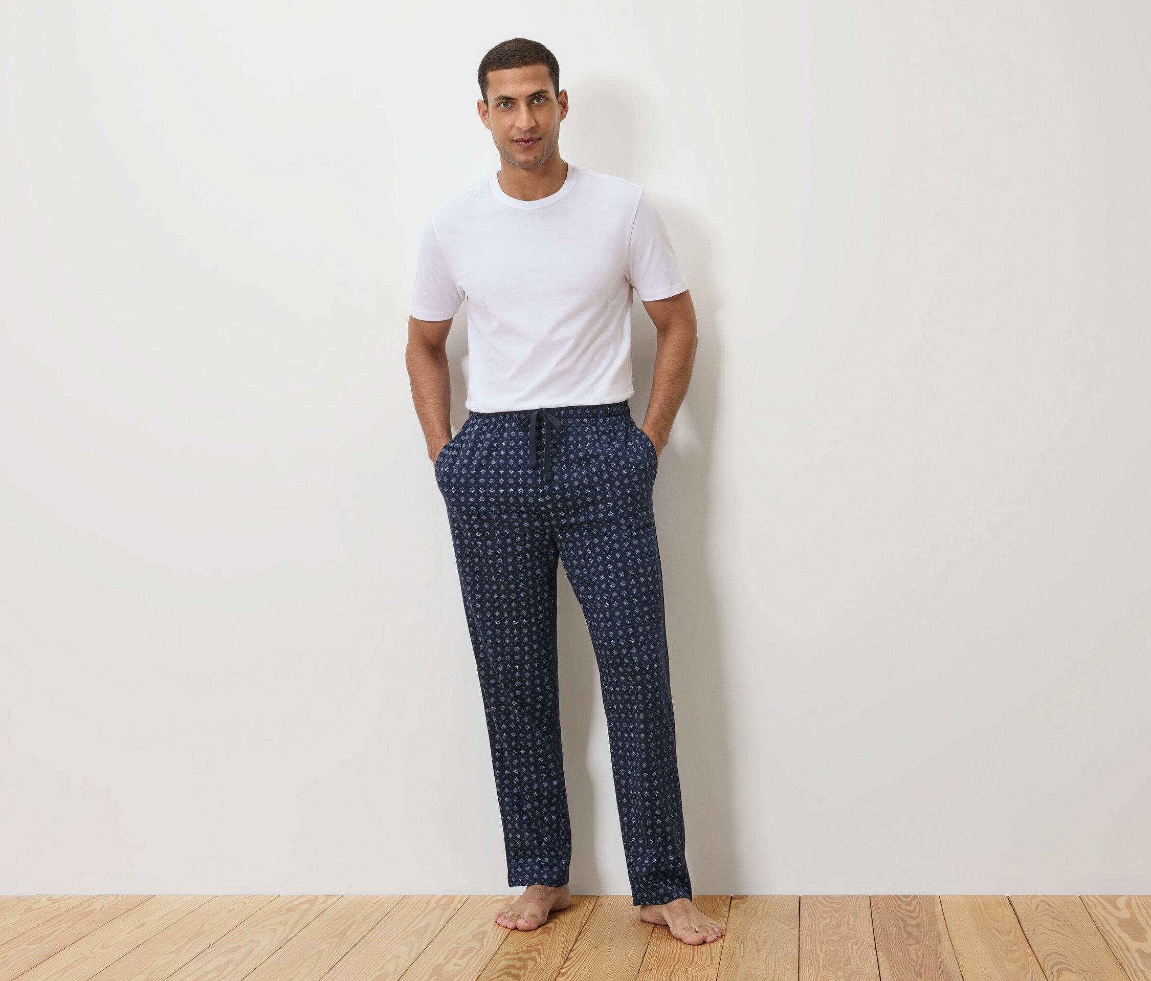 Ein Mann steht vor einer weißen Wand, er trägt ein weißes T-Shirt und eine dunkelblaue Schlafanzughose mit Punktmuster.