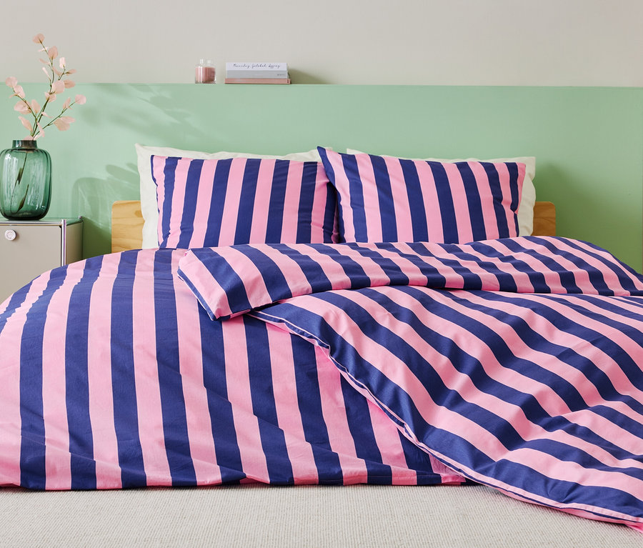 Bett mit blau-rosa gestreifter Bettwäsche, zwei Kissen und grüner Wand im Hintergrund.