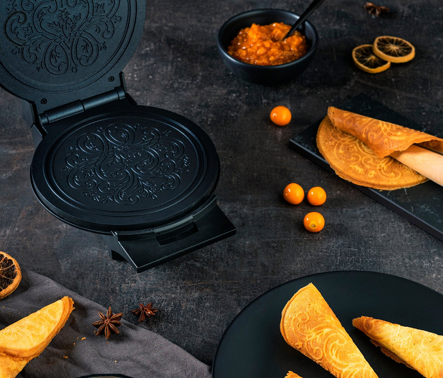Schwarzes WILFA Norwegisches Krumkakeeisen ØSTERDAL Waffeleisen mit gebackenen Krumkaken, einer Schale Obst und Marmelade auf dunklem Hintergrund.
