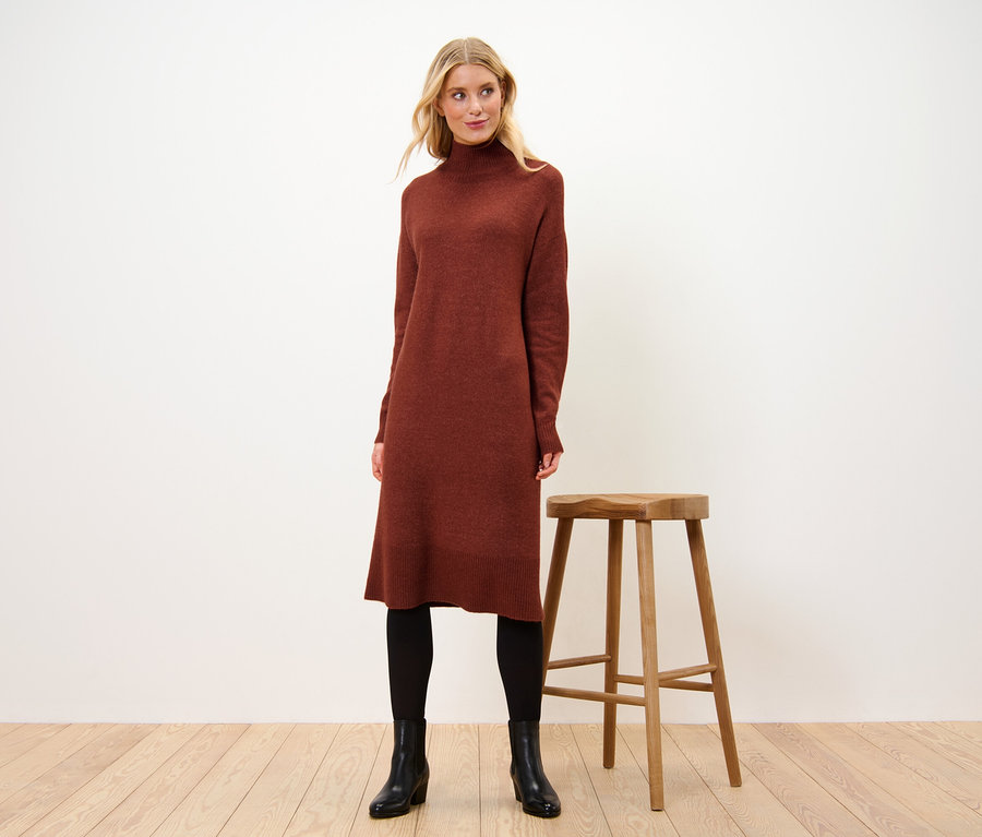 Frau steht neben einem Holzstuhl und trägt ein Strickkleid mit Wollanteil.