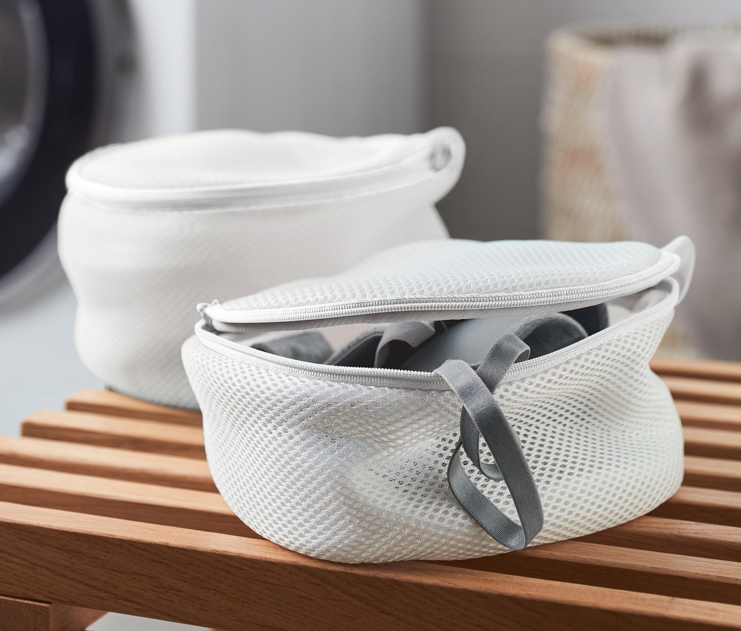 Zwei weiße BH-Wäschenetze auf einer Holzbank, im Hintergrund eine Waschmaschine.