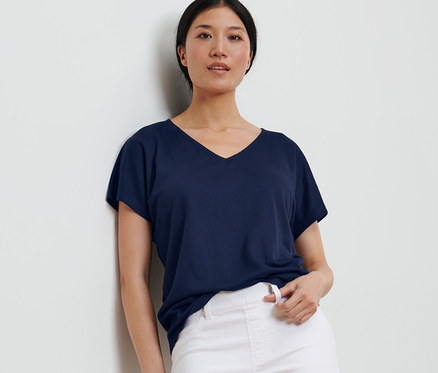 Eine Frau, die sich an eine Wand lehnt, trägt ein dunkelblaues, strukturiertes Shirt und weiße Hosen.