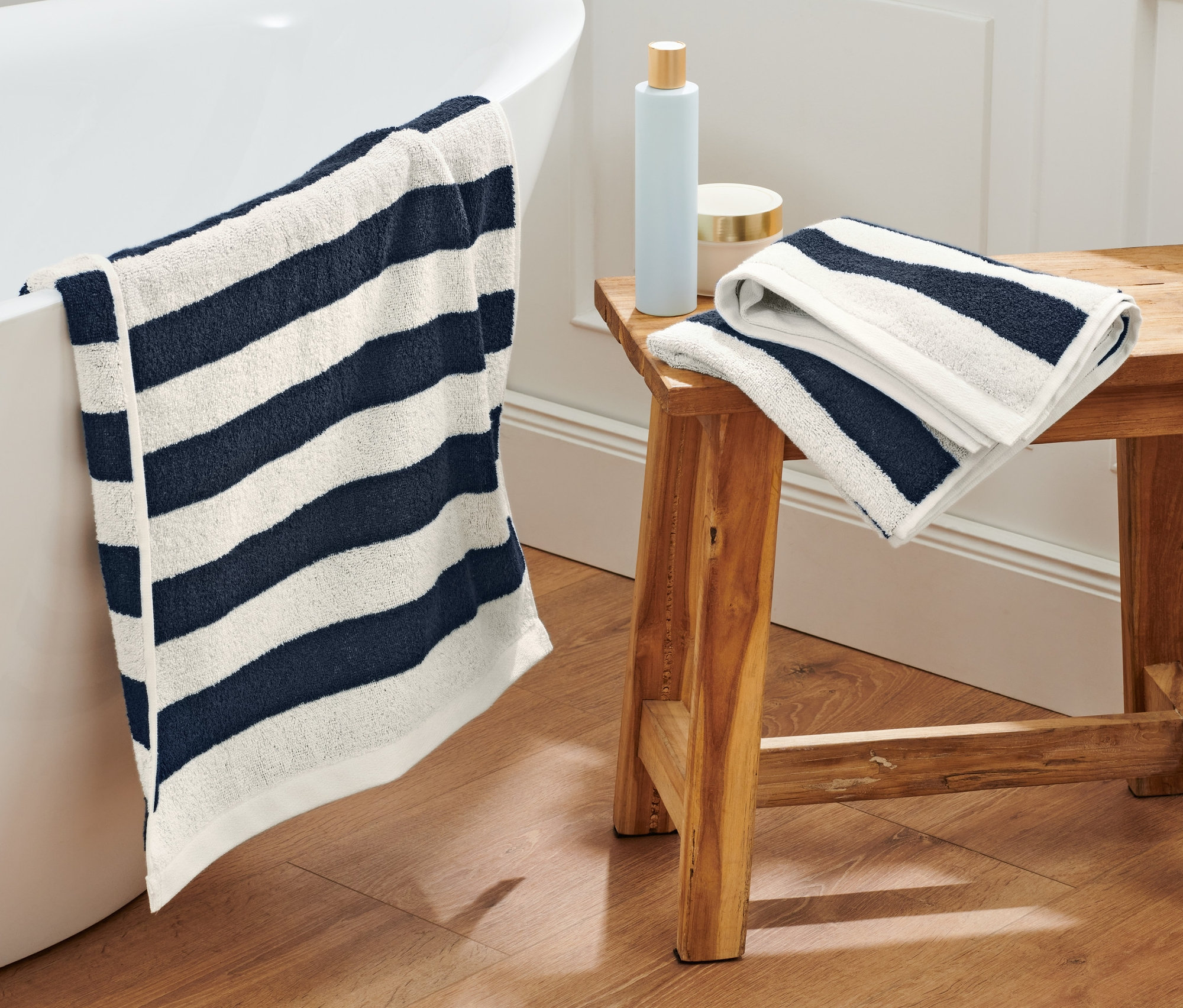 Ein dunkelblau-weiß gestreiftes Handtuch hängt über dem Rand einer Badewanne. Zwei weitere dunkelblau-weiß gestreifte Handtücher liegen auf einem Holzhocker.