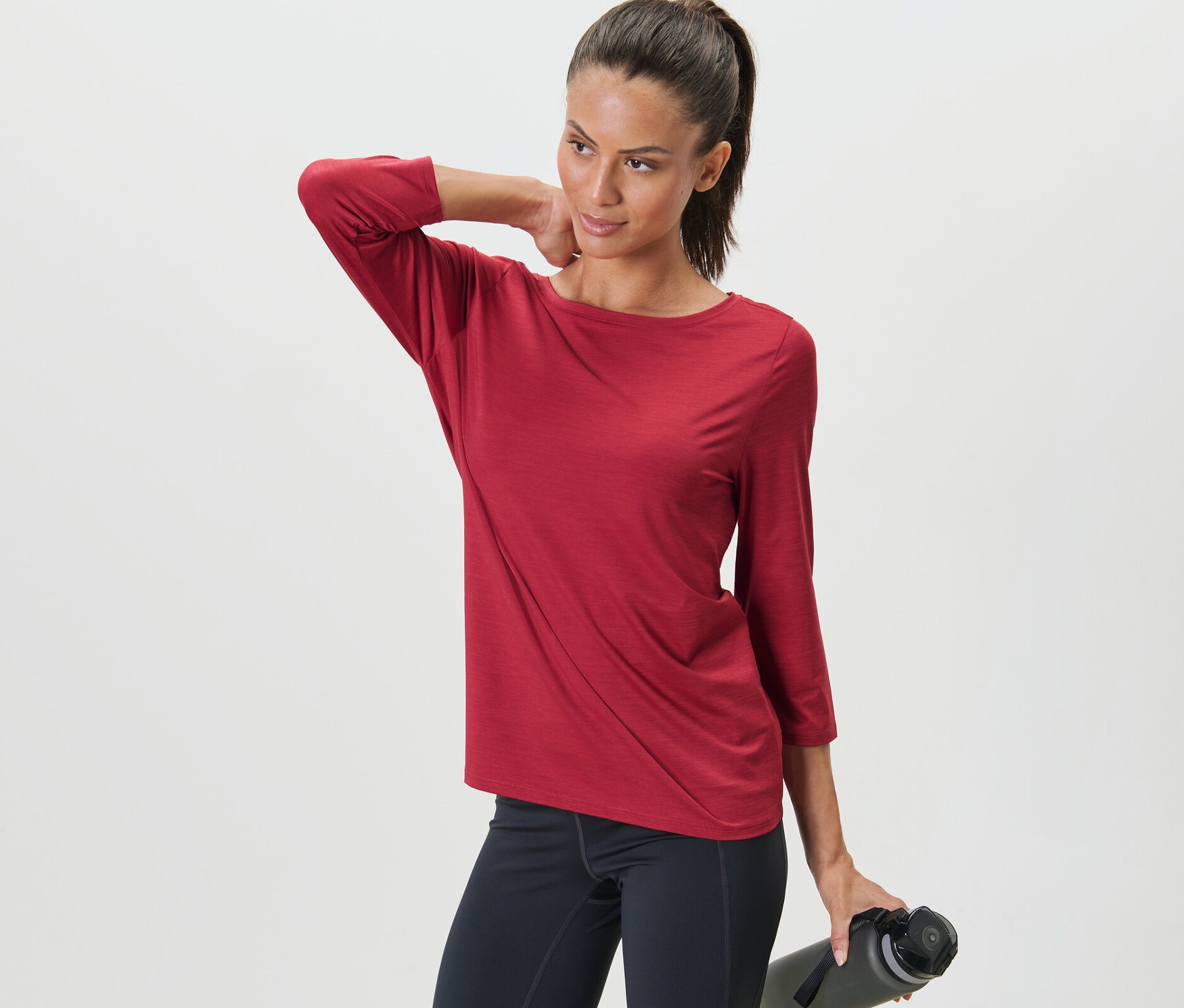 Frau posiert in einem roten Funktionsshirt mit 3/4-Arm und schwarzen Leggings, eine Wasserflasche in der Hand haltend.