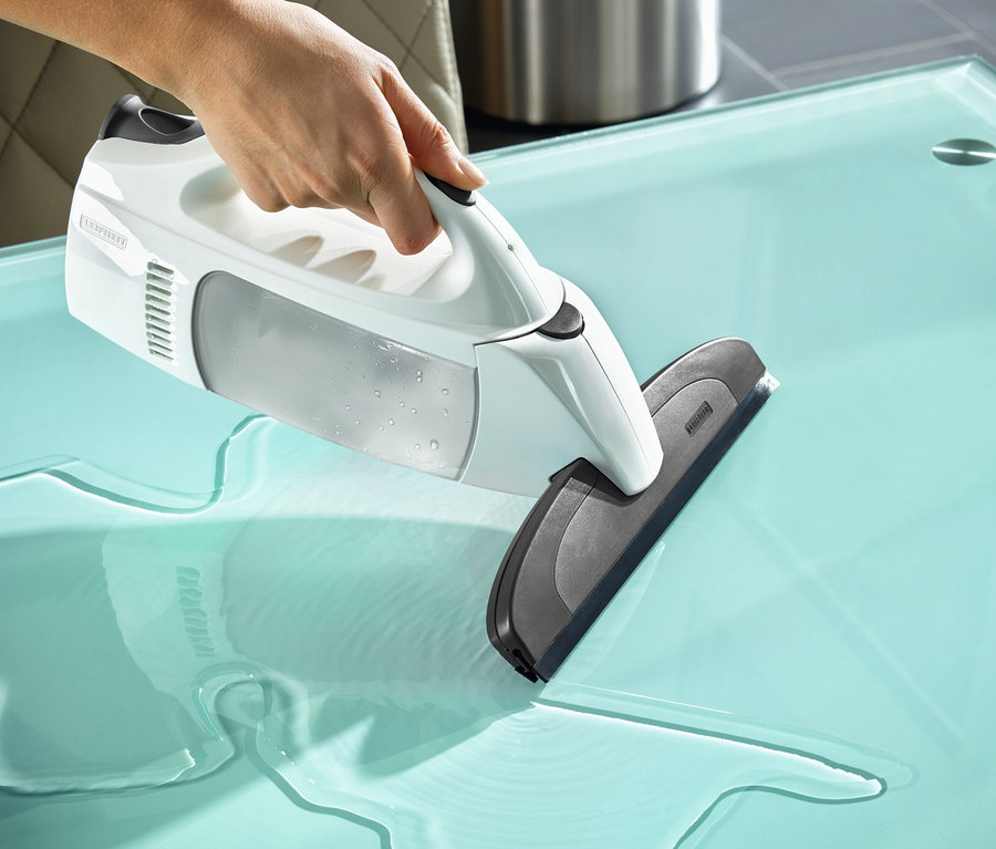 Eine Hand hält einen weißen Fenstersauger »DRY &amp; Clean Classic« auf einer gläsernen Tischplatte mit verschüttetem Wasser.