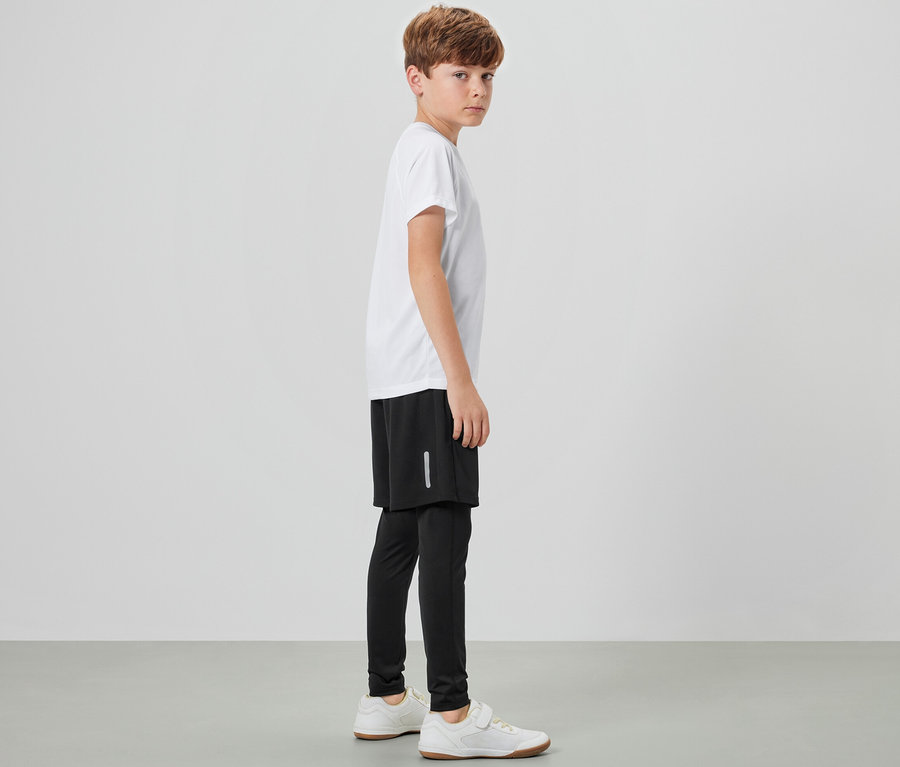 Ein Junge steht seitlich und trägt eine Kinder-Sporttight mit Shorts, ein weißes T-Shirt und Turnschuhe.