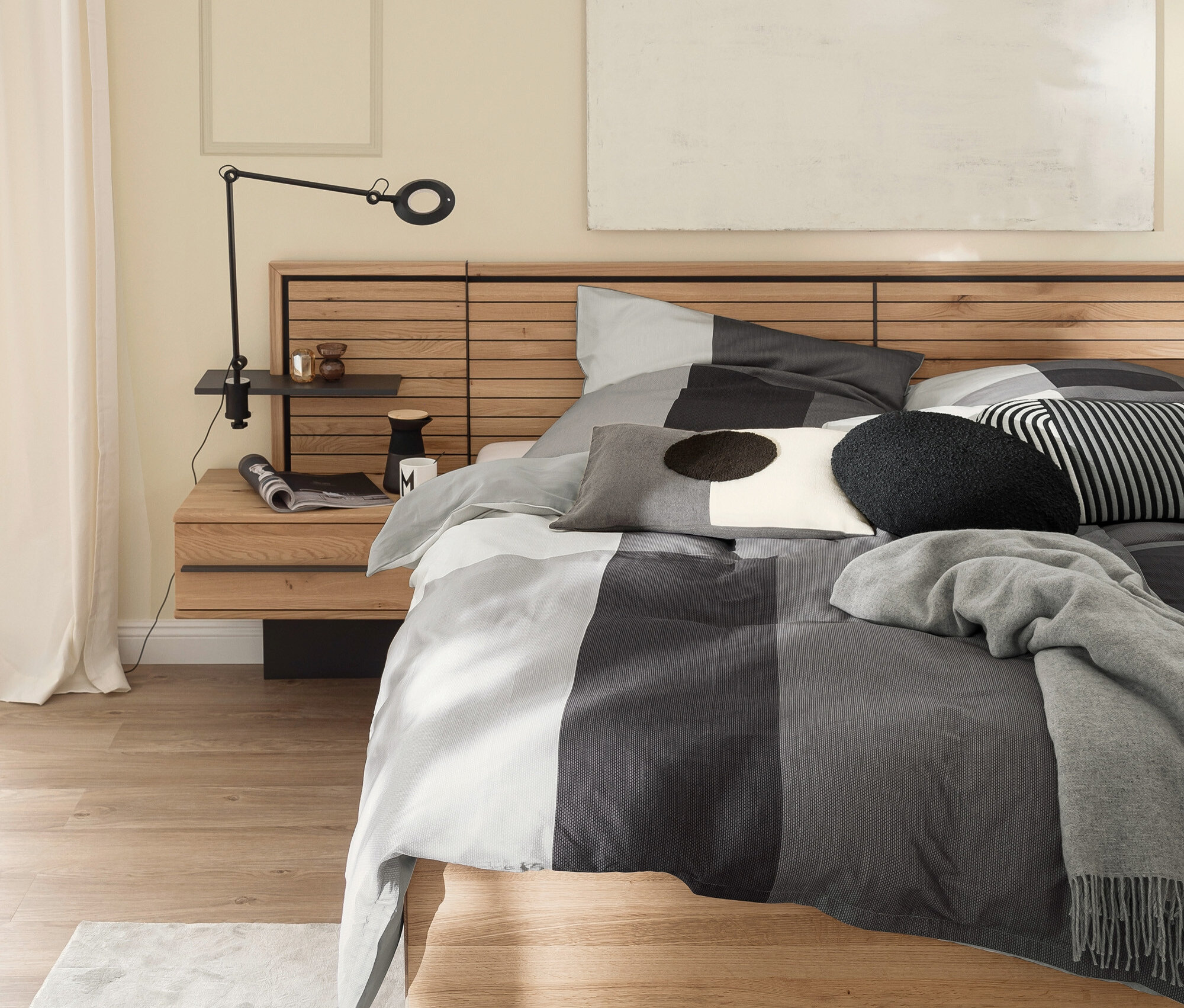 Bett mit Holzkopfteil, grau-weiß gestreifter Bettwäsche und Lampe auf dem Nachttisch.