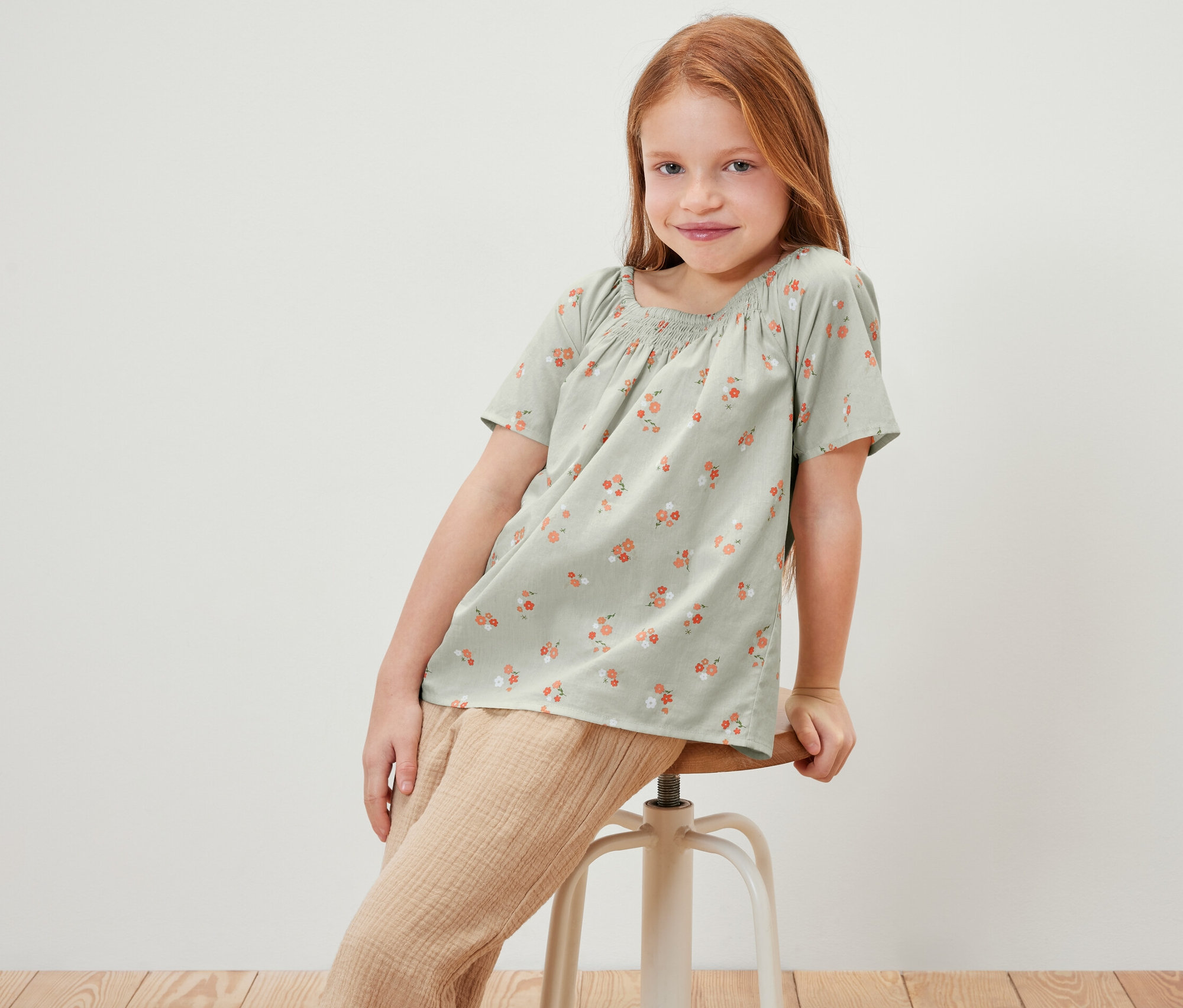 Mädchen sitzt auf einem Hocker und trägt eine Kinder-Bluse mit Blumenmuster.
