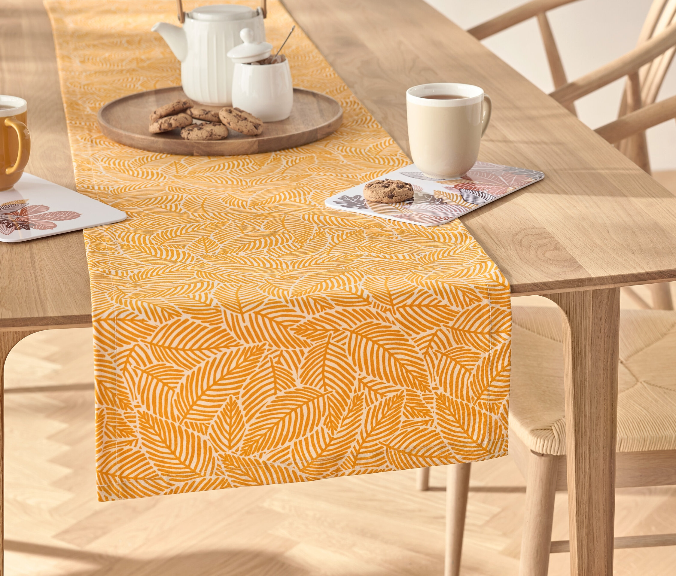 Auf einem Holztisch liegt ein gelber Tischläufer mit Blattmuster, auf dem sich ein Teeservice, eine Tasse und Kekse befinden.