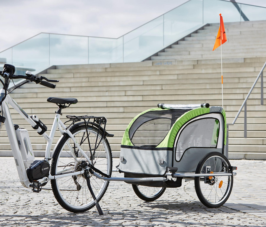 Weißes E-Bike mit montiertem FISCHER Kinder-Komfort-Fahrradanhänger vor einer Treppe.