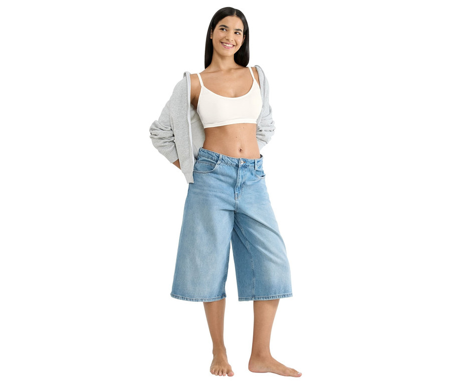 Eine barfüßige Frau steht da und trägt ein weißes Crop-Top, weite Jeansshorts und eine graue Jacke.