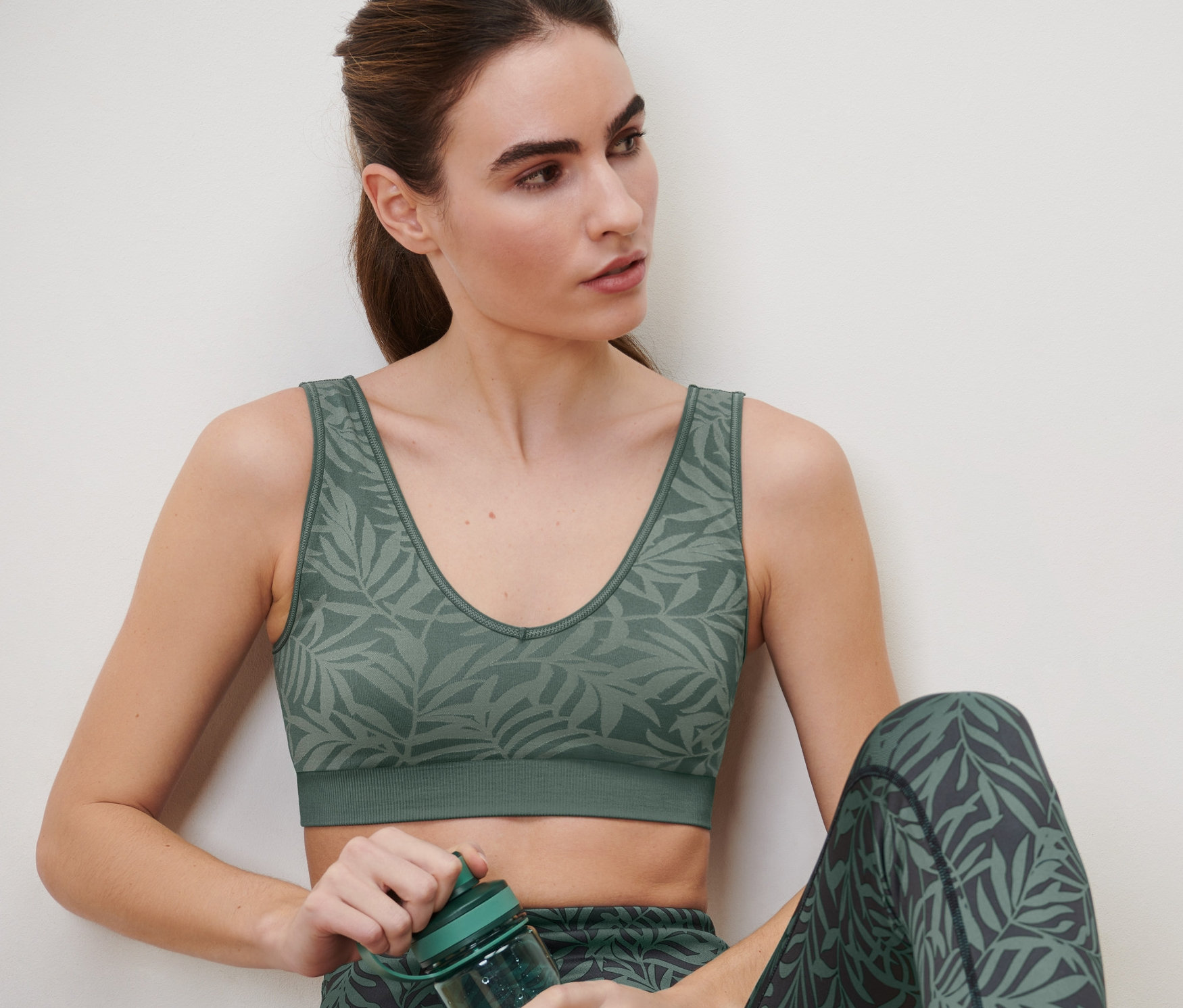 Eine Frau sitzt und hält eine grüne Sportflasche. Sie trägt ein Seamless-Sport-Bustier in Salbei und Leggings mit Blätterdruck.