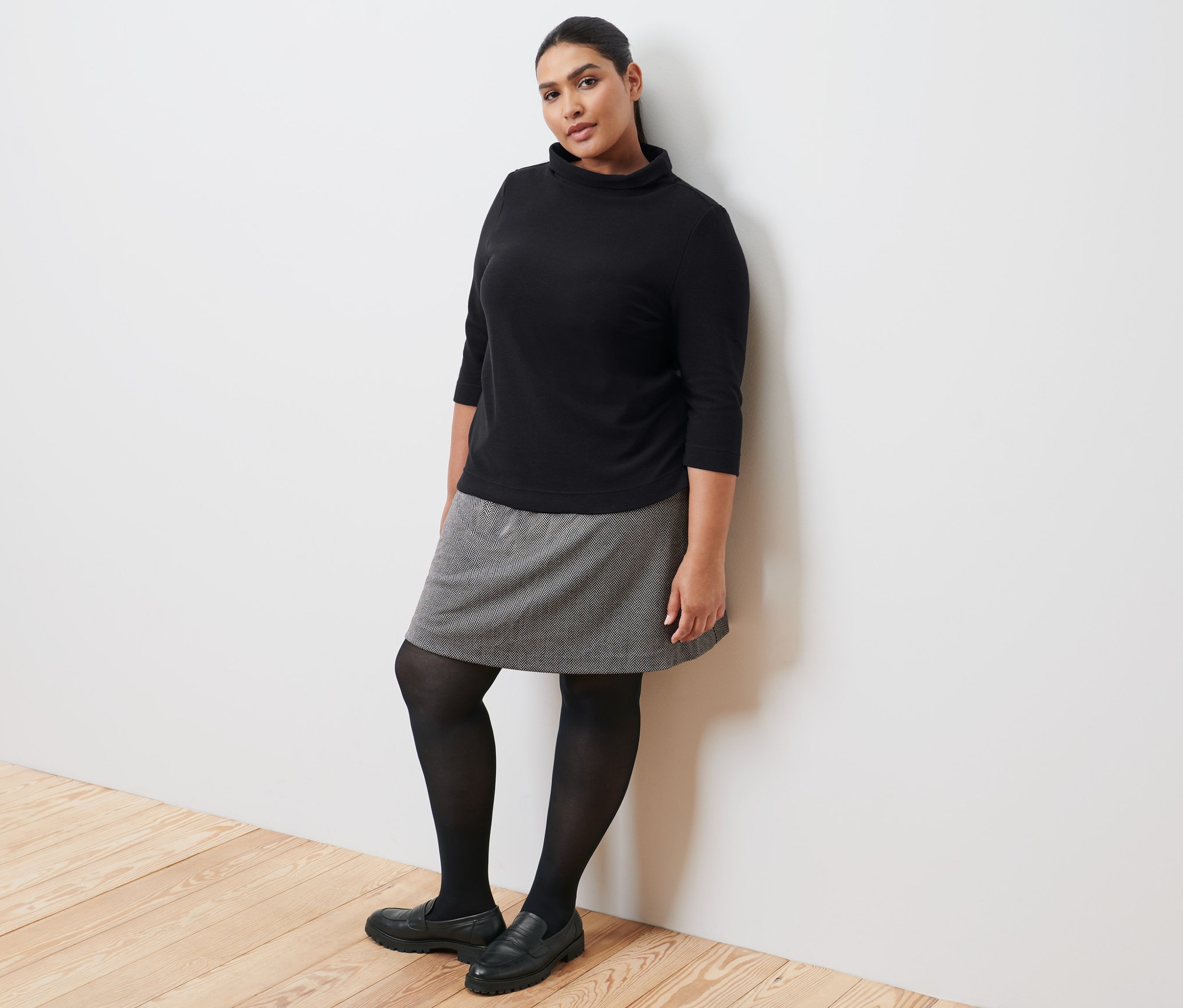 Eine Frau, die an eine Wand gelehnt ist, trägt ein schwarzes Sweatshirt mit 3/4-Arm, einen Sweatrock, eine schwarze Strumpfhose und Leder-Loafer.