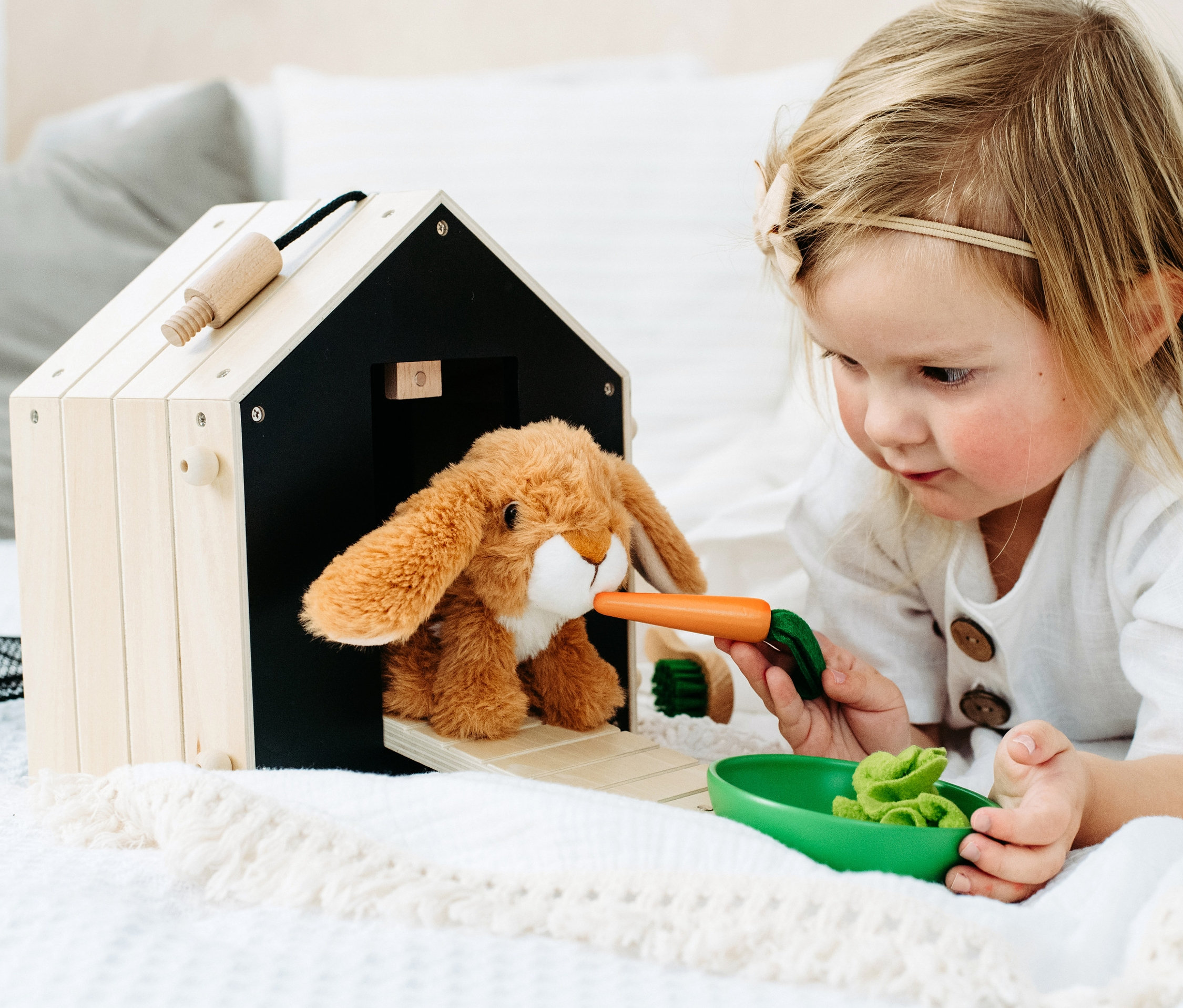 Mädchen füttert Plüschhasen mit Karotte vor small foot Hasenstall mit Auslauf.