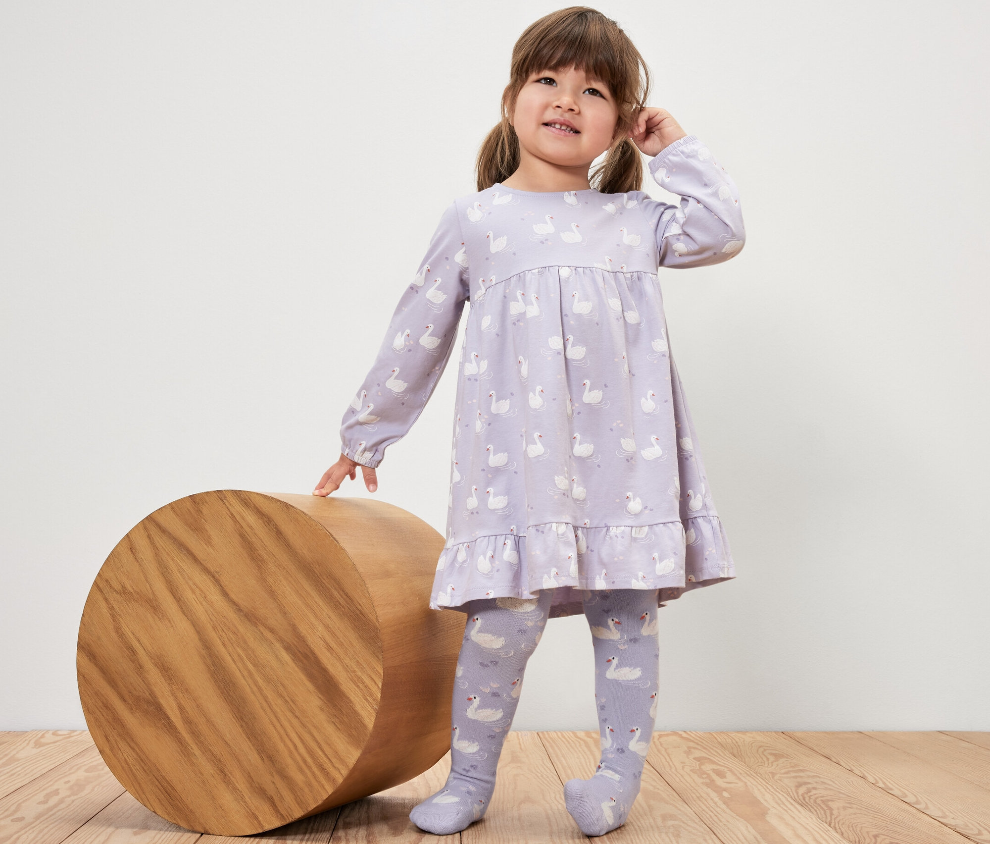 Mädchen im Kleid mit Schwanenmuster steht neben einem Holzzylinder und trägt 5 Kinder-Strumpfhosen, Prinzessin.