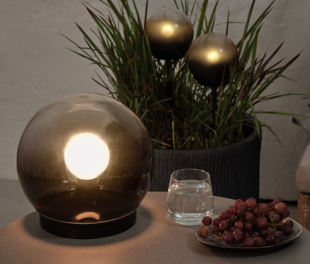 Auf einem Tisch stehen eine Rauchglas-Kugelleuchte, ein Glas Wasser und Trauben. Im Hintergrund befinden sich 2 LED-Kugelleuchten in einem Blumentopf.