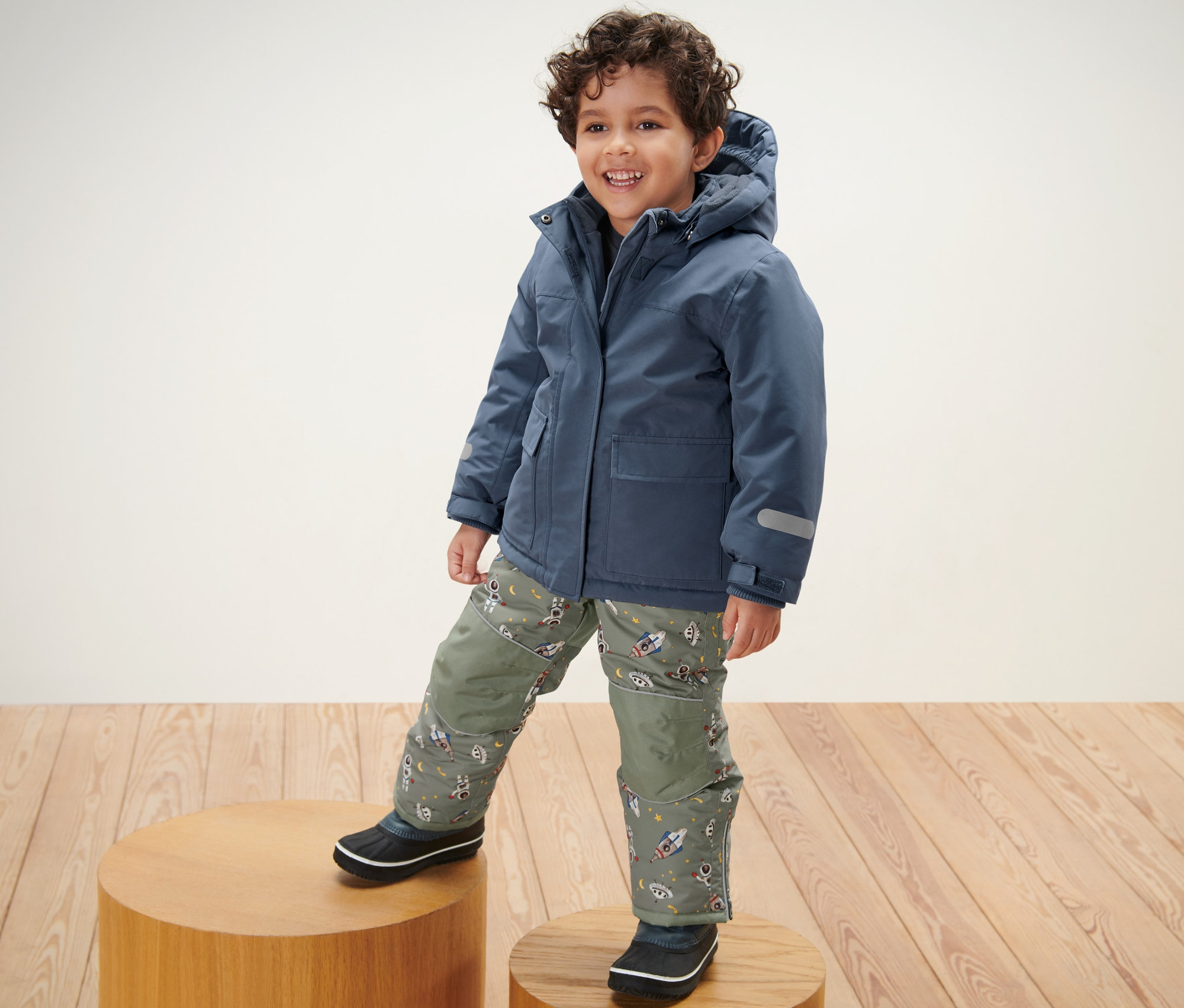 Junge steht auf einem Holzpodest und trägt eine Kinder-Schneekombi.