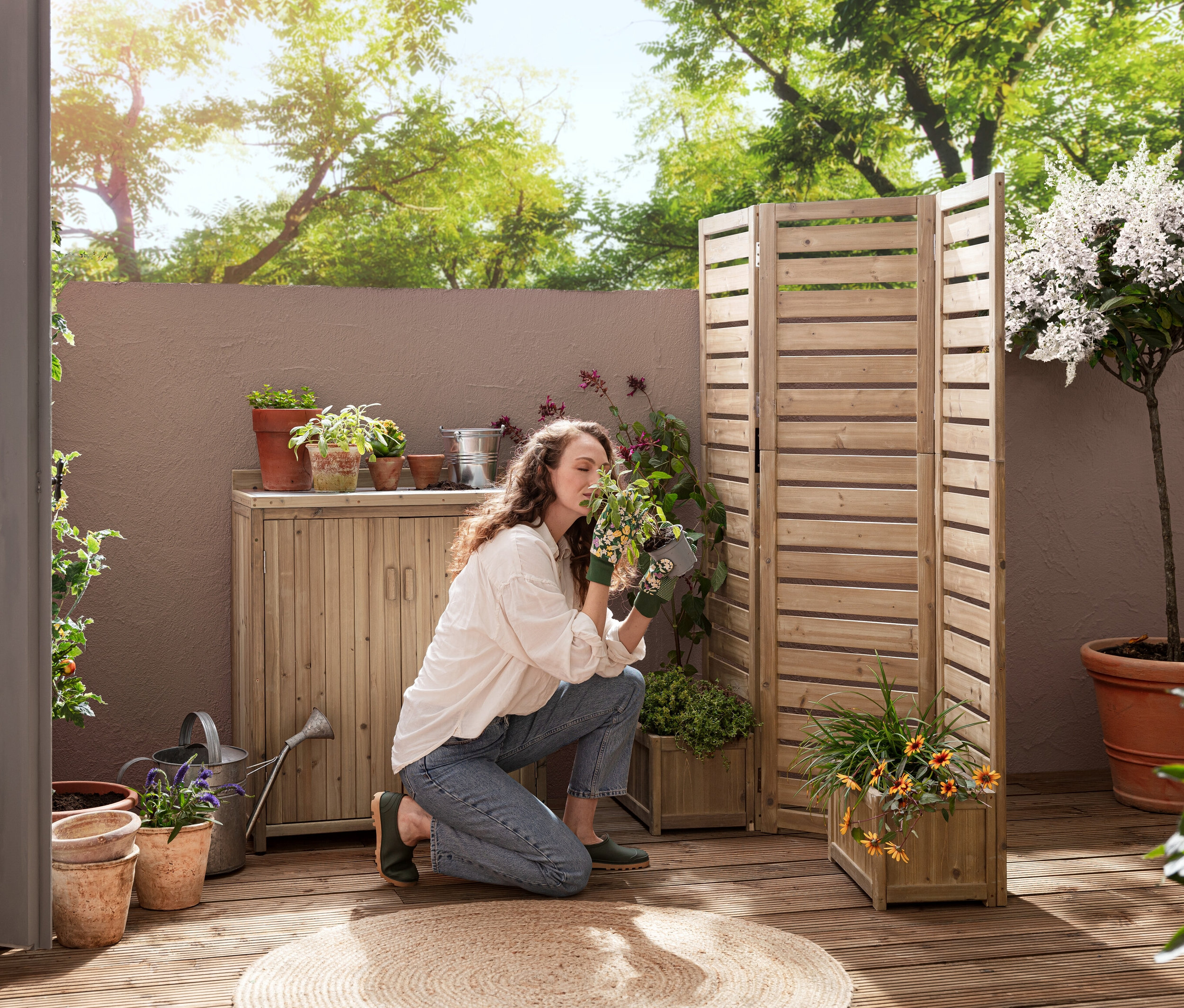 Eine Frau kniet auf einer Terrasse und riecht an einer Pflanze in der Nähe eines flexiblen Paravents »Tinus« mit Pflanzkästen und eines kleinen Gartenschranks »Tinus«.