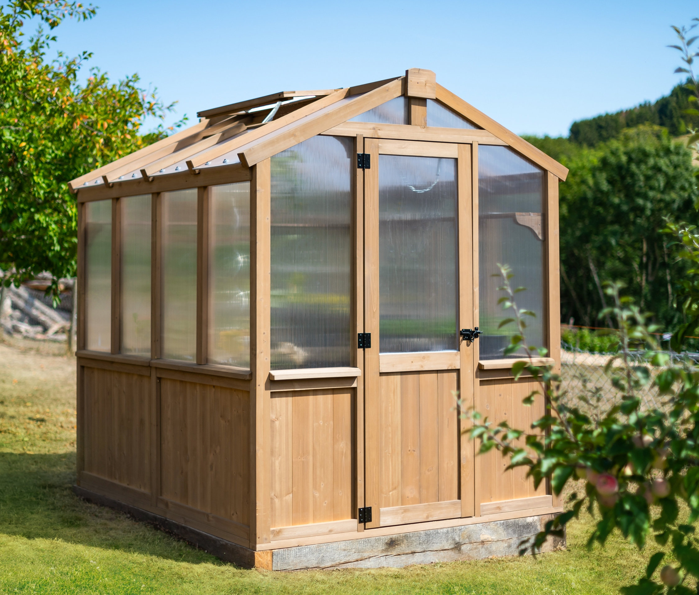 Ein Gewächshaus aus Zedernholz steht an einem sonnigen Tag auf einem Rasen.
