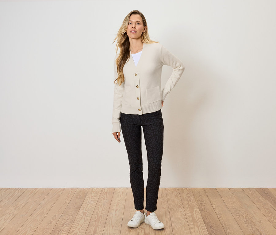 Blonde Frau posiert in cremefarbenem Cardigan, gemusterter, schwarzer Hose und weißen Turnschuhen.