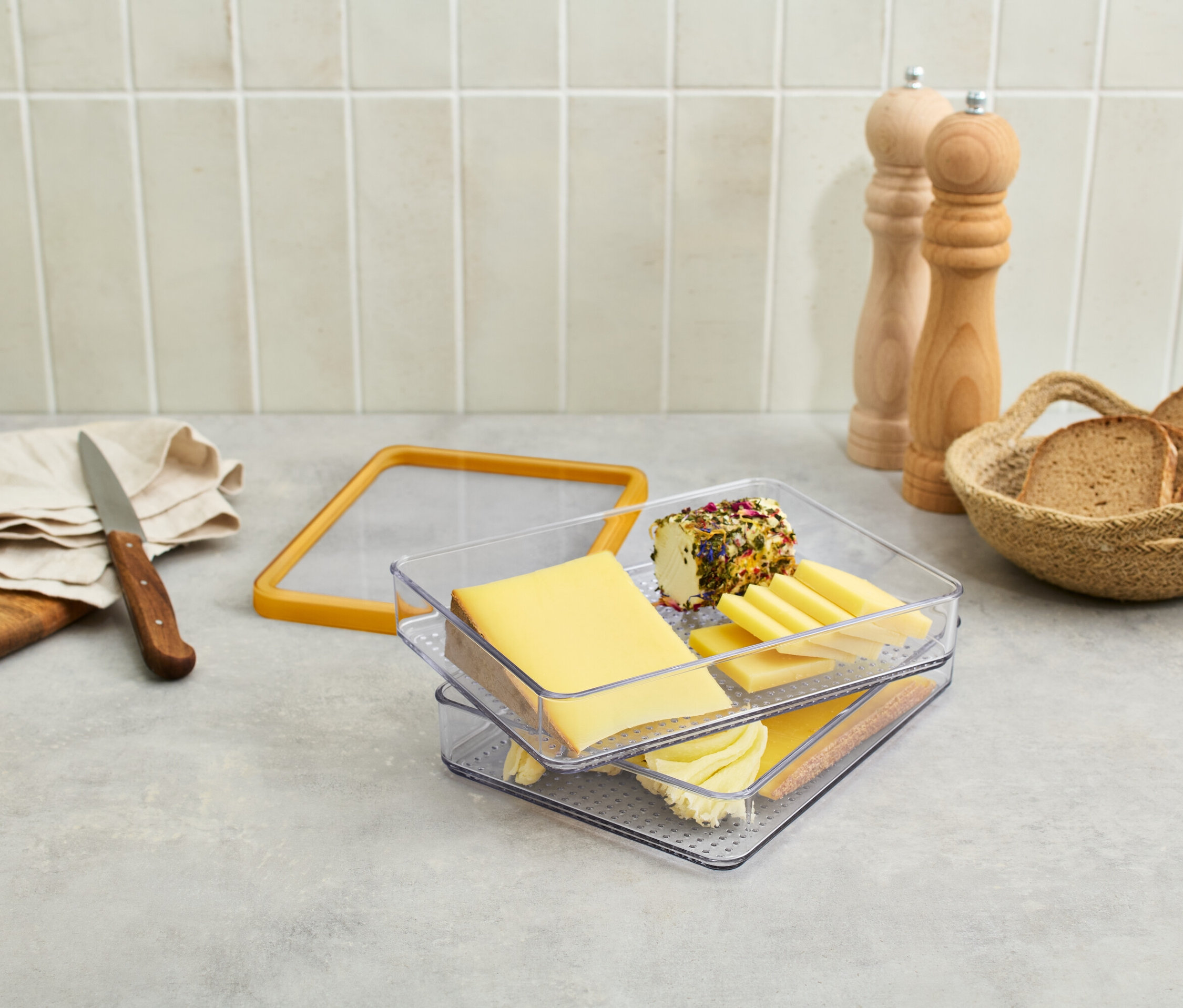 Auf einem Tisch stehen Käse in einem durchsichtigen Behälter, Trauben, ein Messer, Brot in einem Korb und Gewürzmühlen.