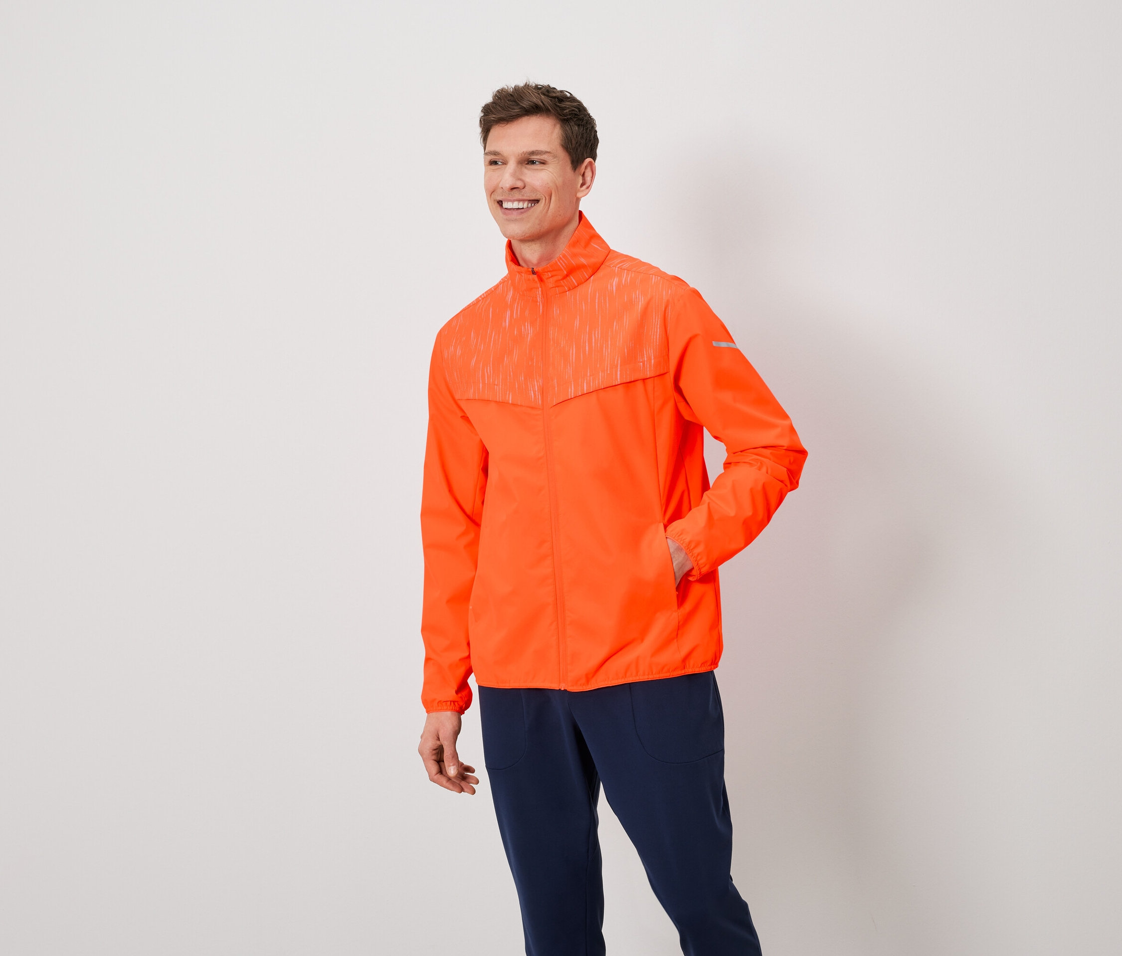 Ein Mann mit braunen Haaren trägt eine orangefarbene Jacke und dunkelblaue Hosen.