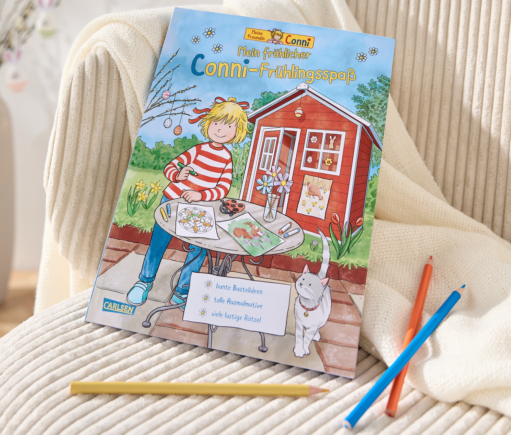 Buch »Mein fröhlicher Conni-Frühlingsspaß« liegt auf einem cremefarbenen Sessel neben Buntstiften.