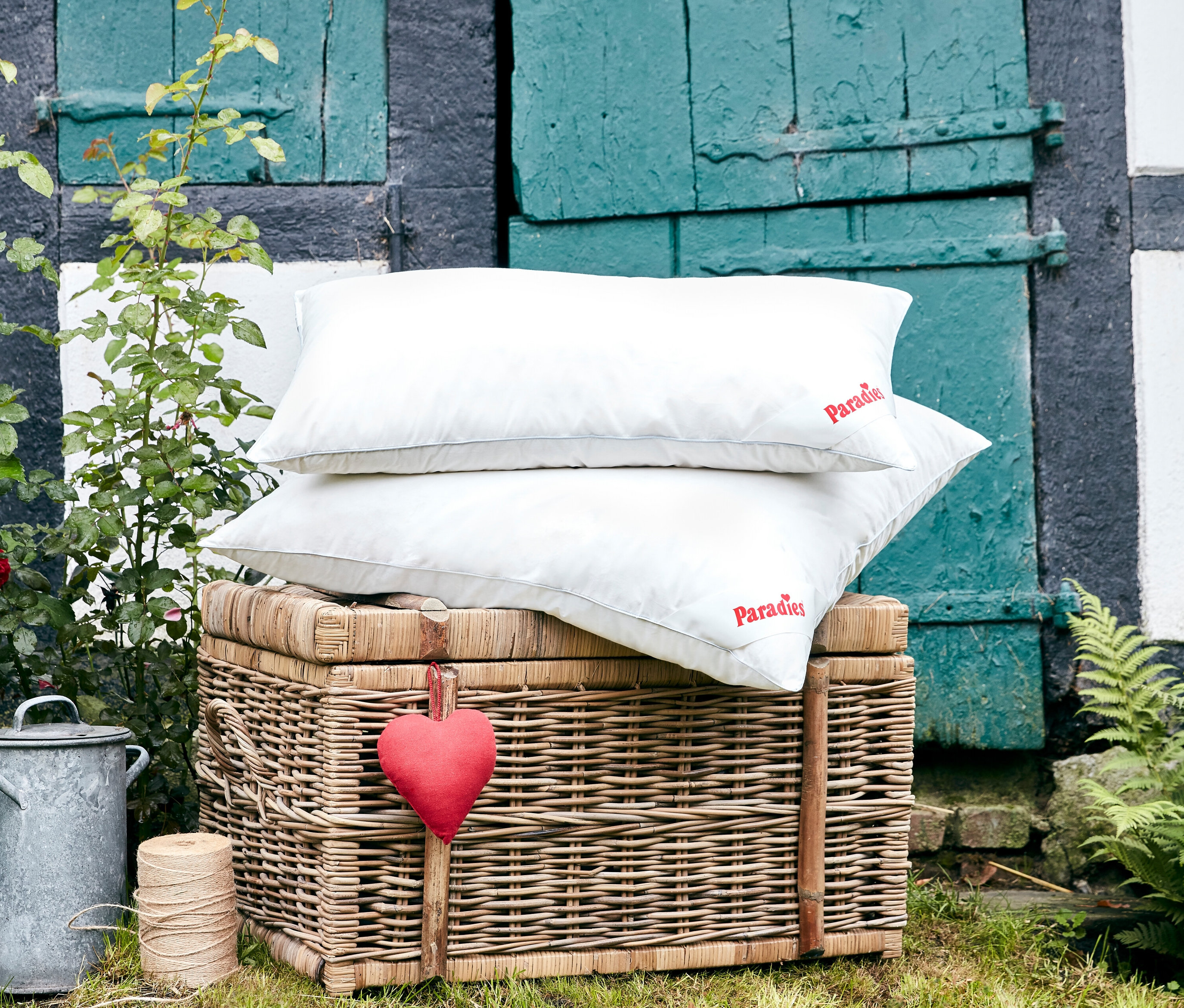 Zwei weiße Paradies »Softy BestCool« Kopfkissen (ca. 80 x 80 cm und 80 x 40 cm) liegen gestapelt auf einem Weidenkorb vor einer blaugrünen Holztür.